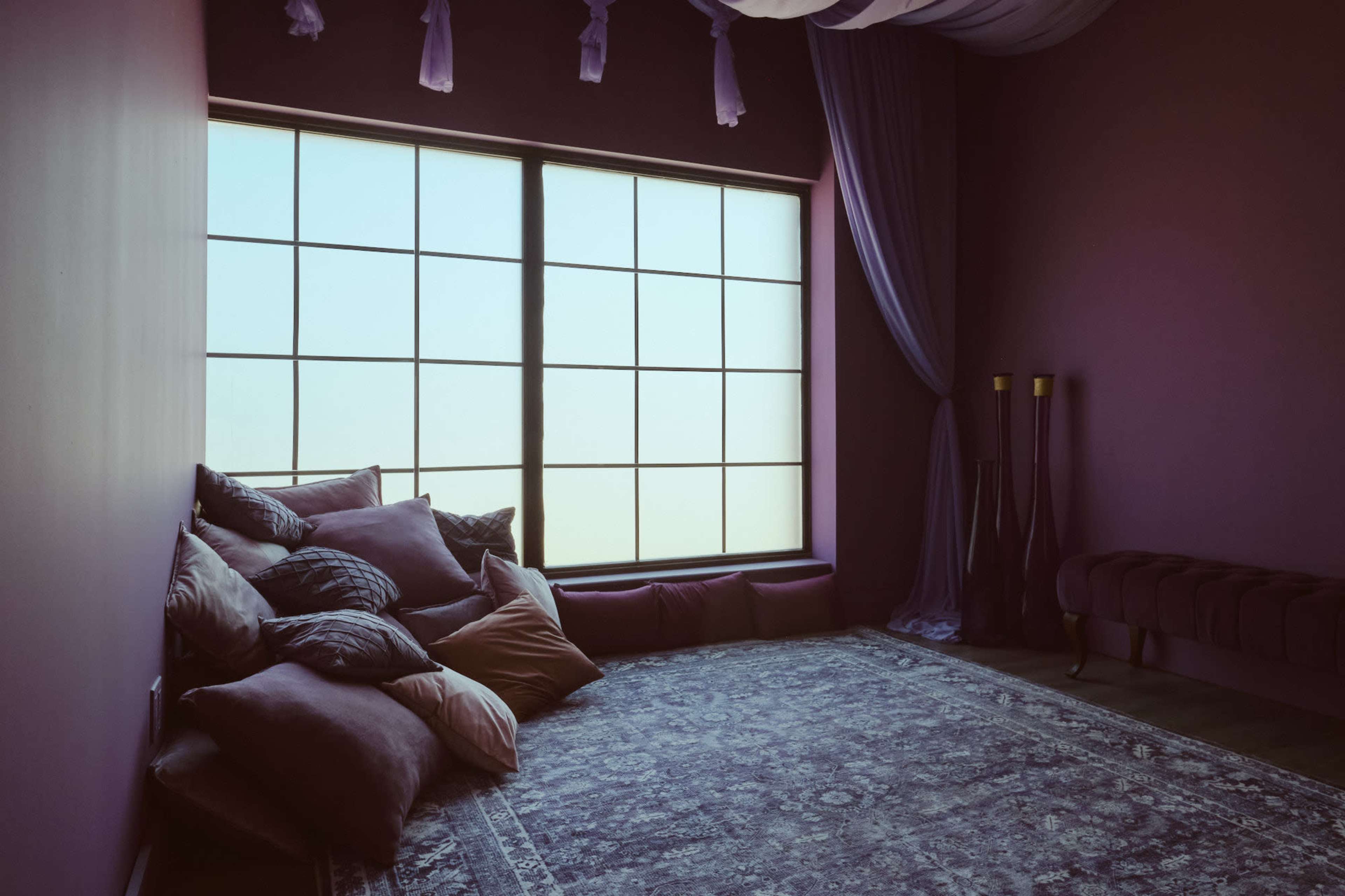 A spacious, purple-themed room features a large window with a grid design and a cozy seating area with plush pillows and a decorative rug.