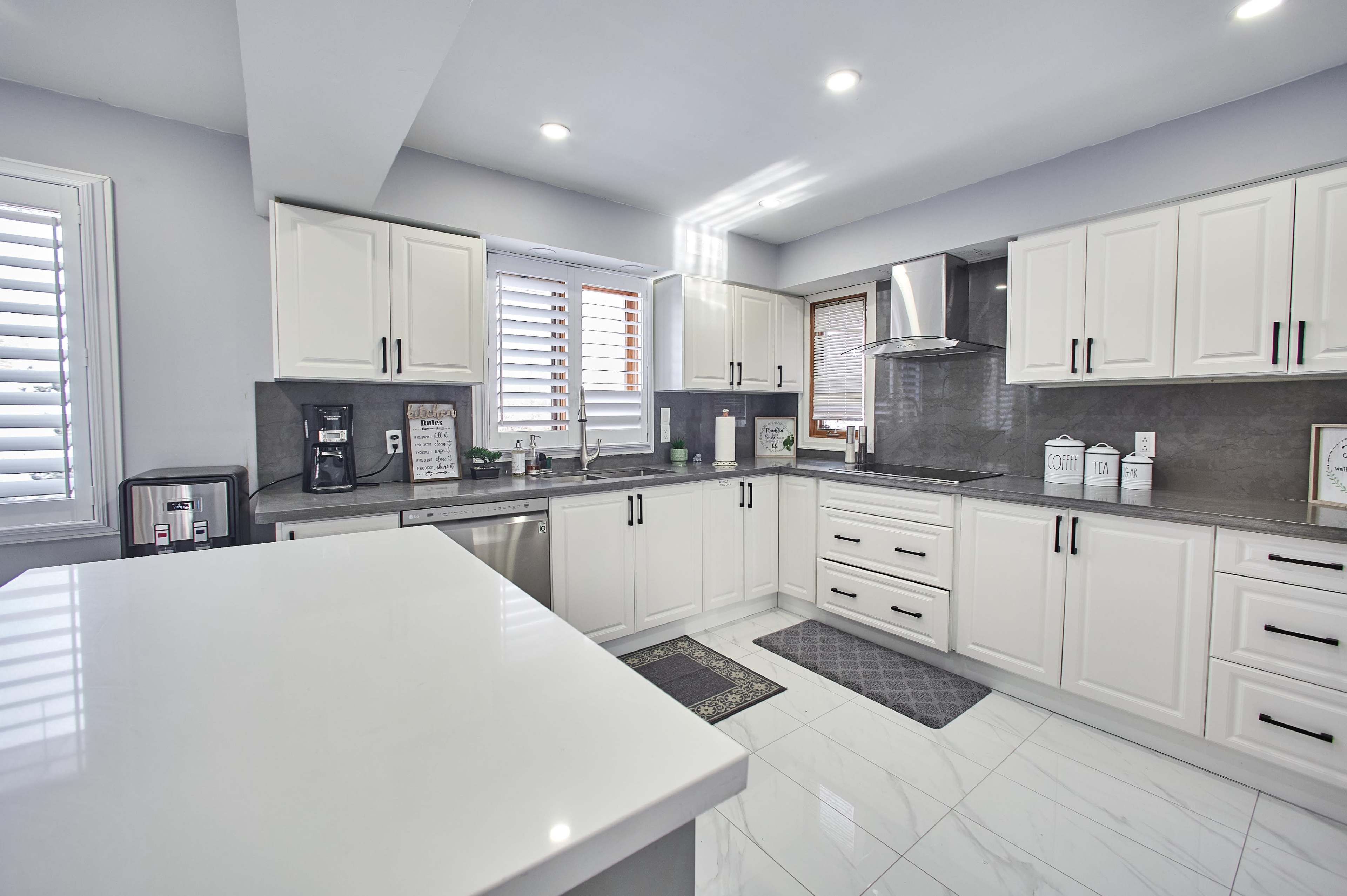 A modern kitchen features white cabinetry, gray countertops, and stainless steel appliances, with ample natural light coming through the windows.