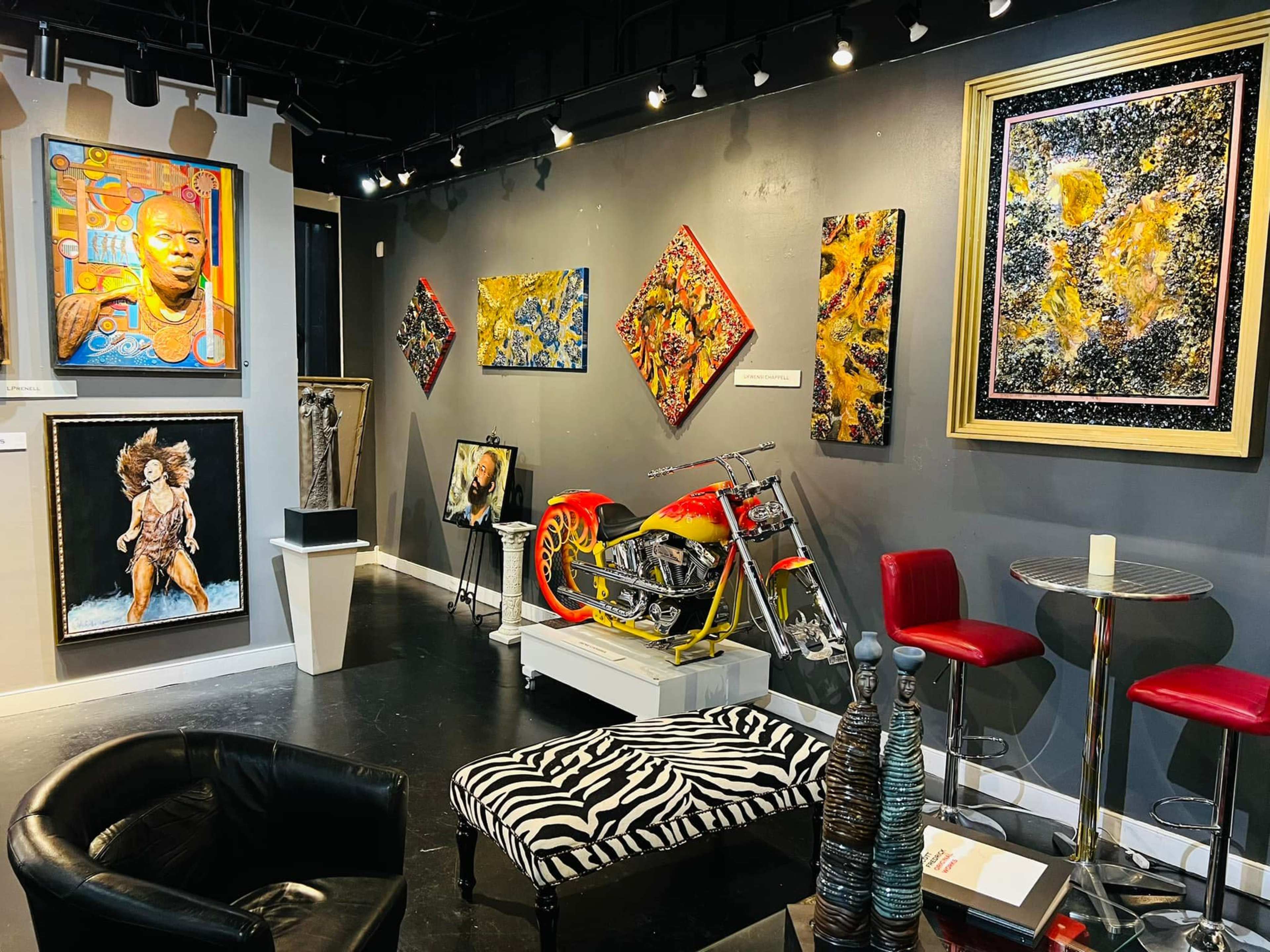 An art gallery with vibrant paintings on the walls, a motorcycle displayed in the center, and a striped ottoman in front of a seating area.