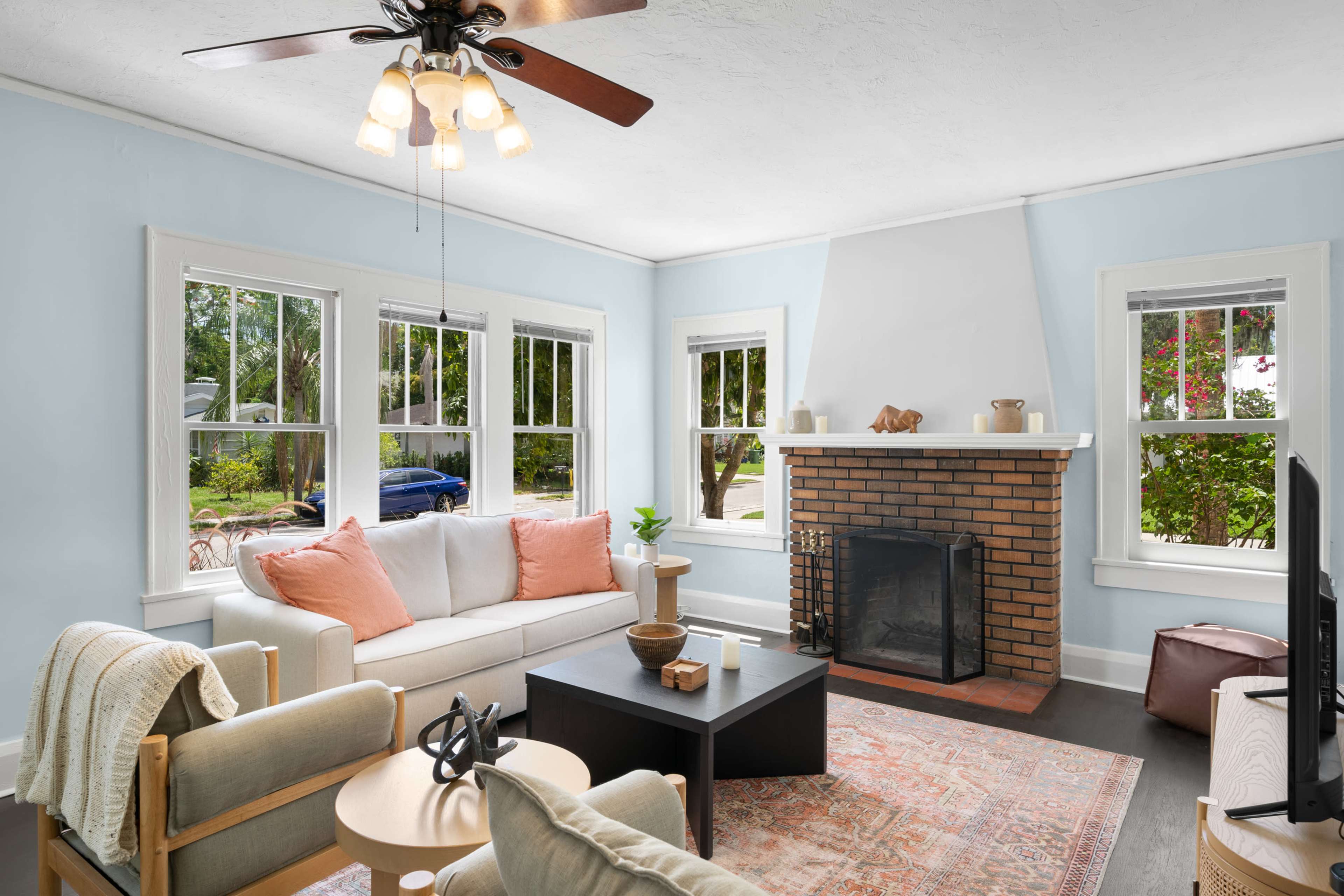 A cozy living room with a light blue wall, a brick fireplace, large windows, and a mix of furniture including a sofa with orange cushions and a black coffee table.