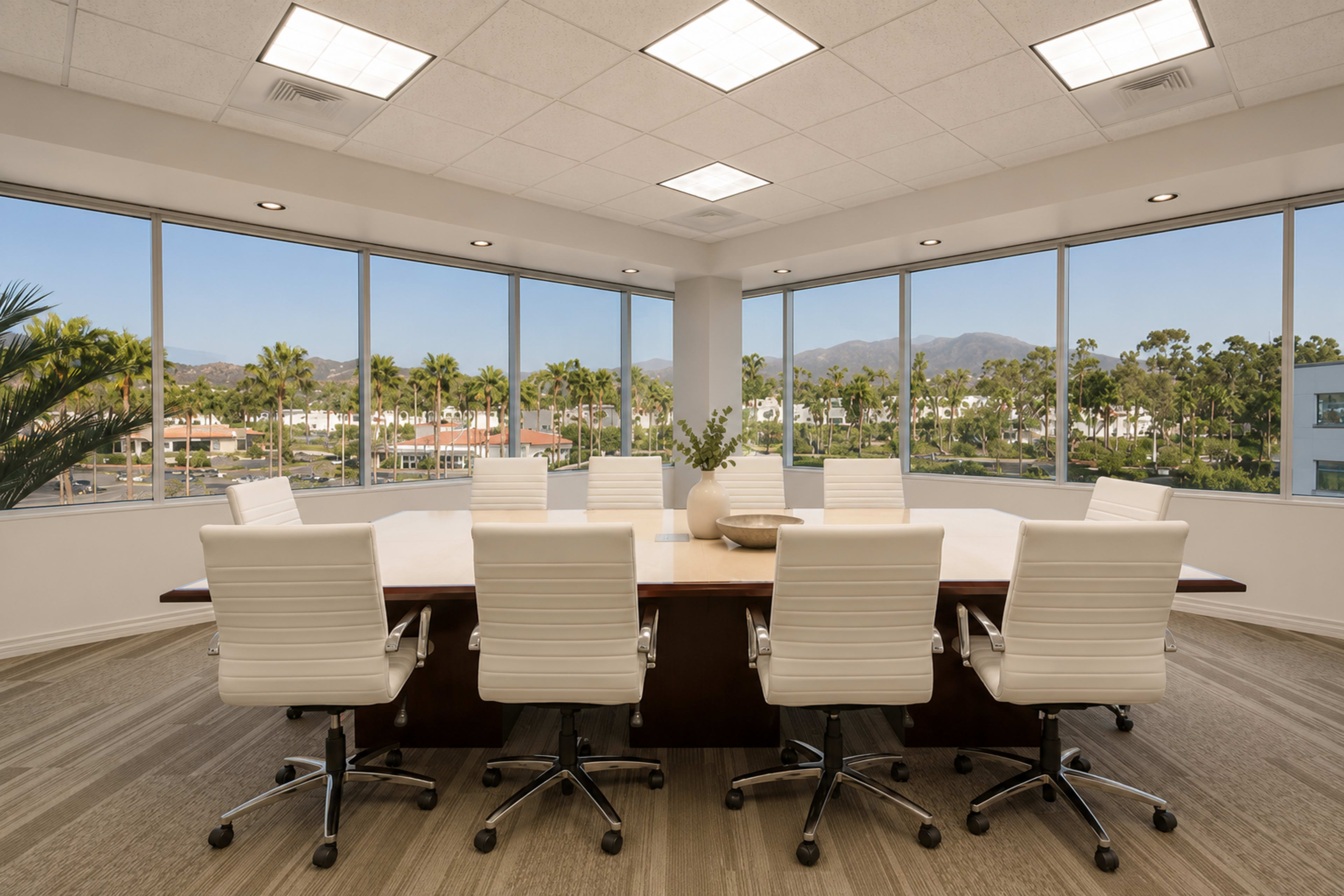 A modern conference room features a large table surrounded by white chairs, with panoramic windows offering a view of palm trees and a distant mountain.