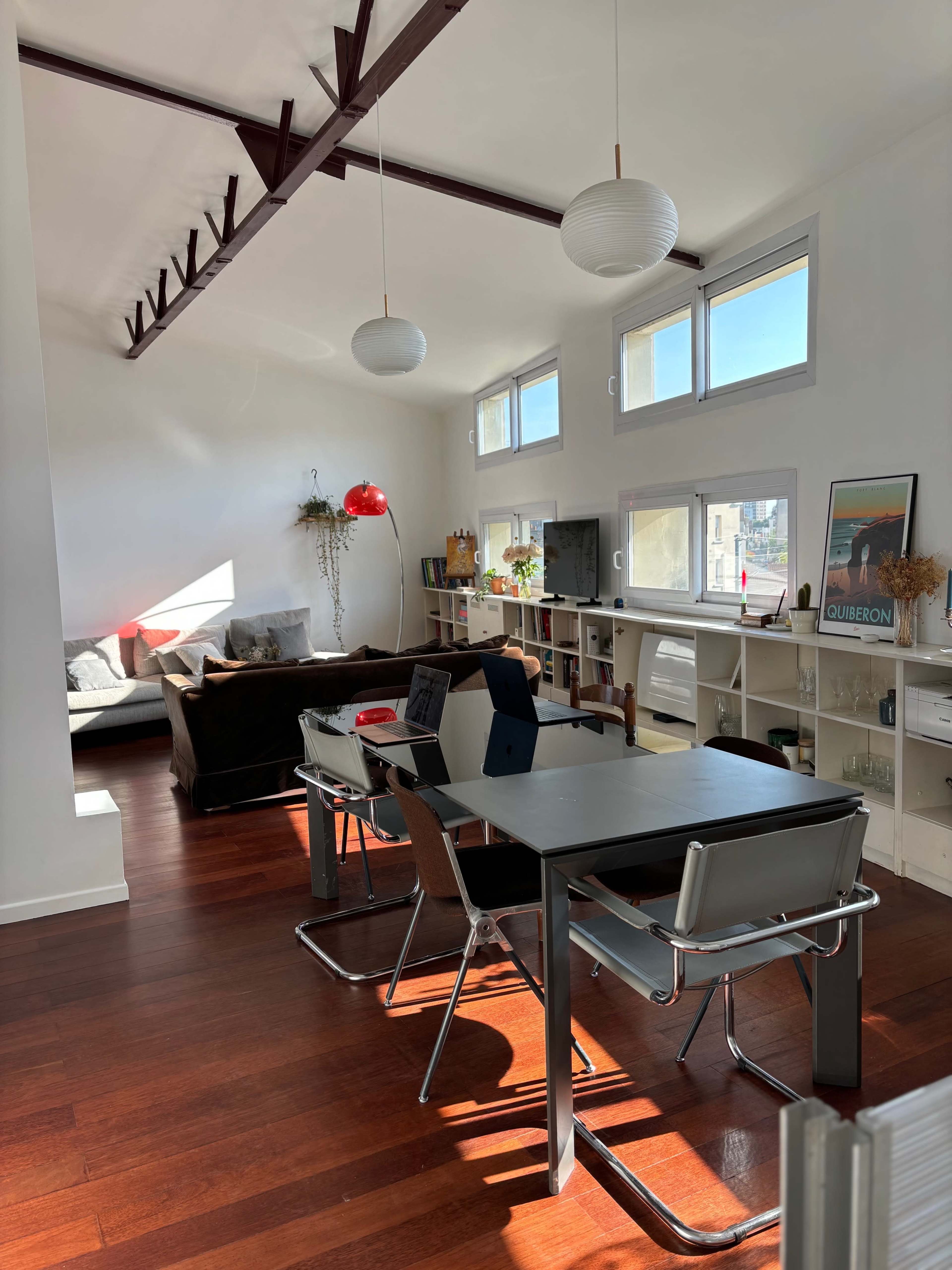 The image shows a bright, modern living space featuring a dining table with chairs, a sofa area, and large windows allowing natural light to enter.