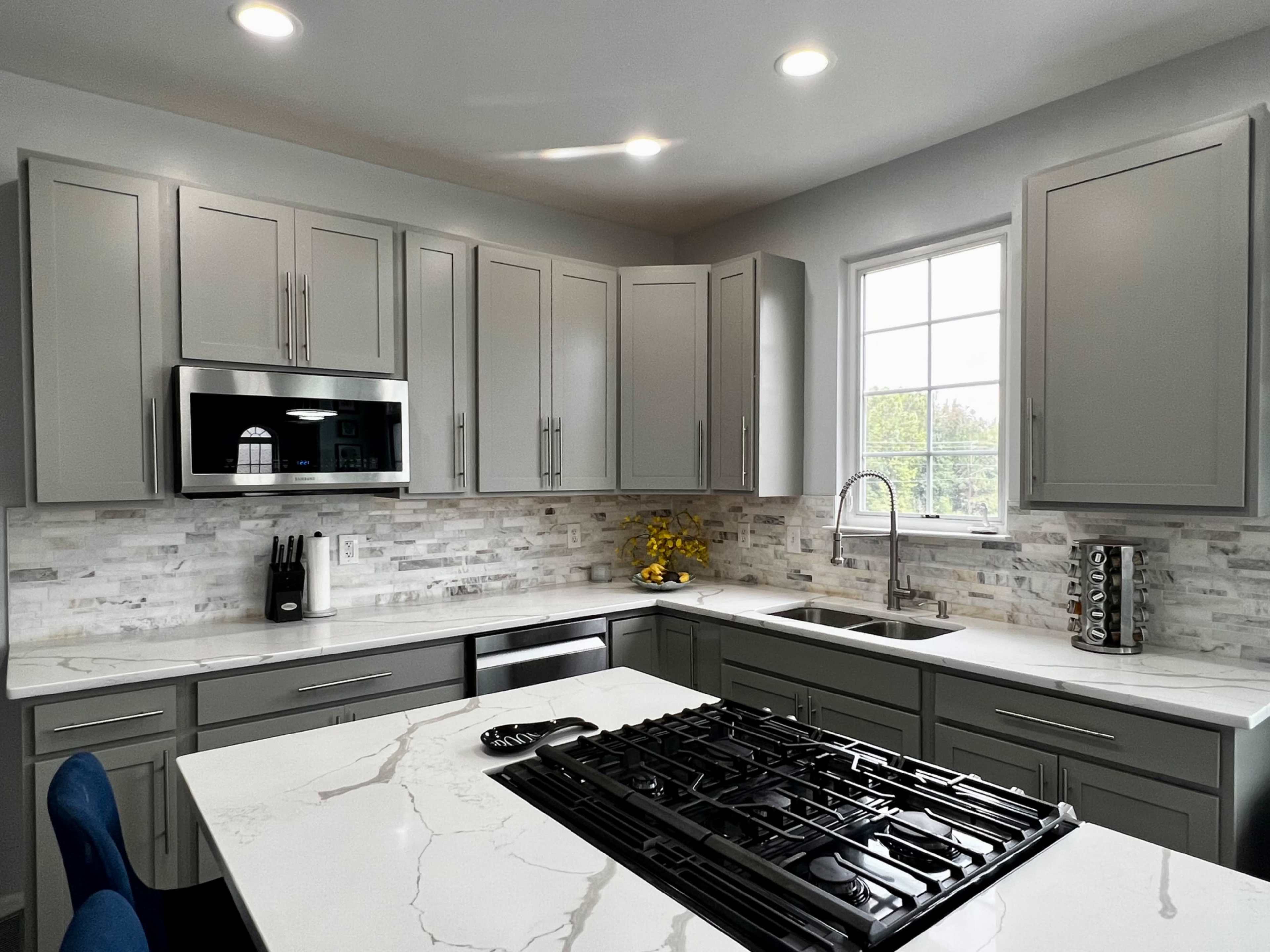 A modern kitchen features gray cabinets, a center island with a gas cooktop, and a window overlooking greenery.