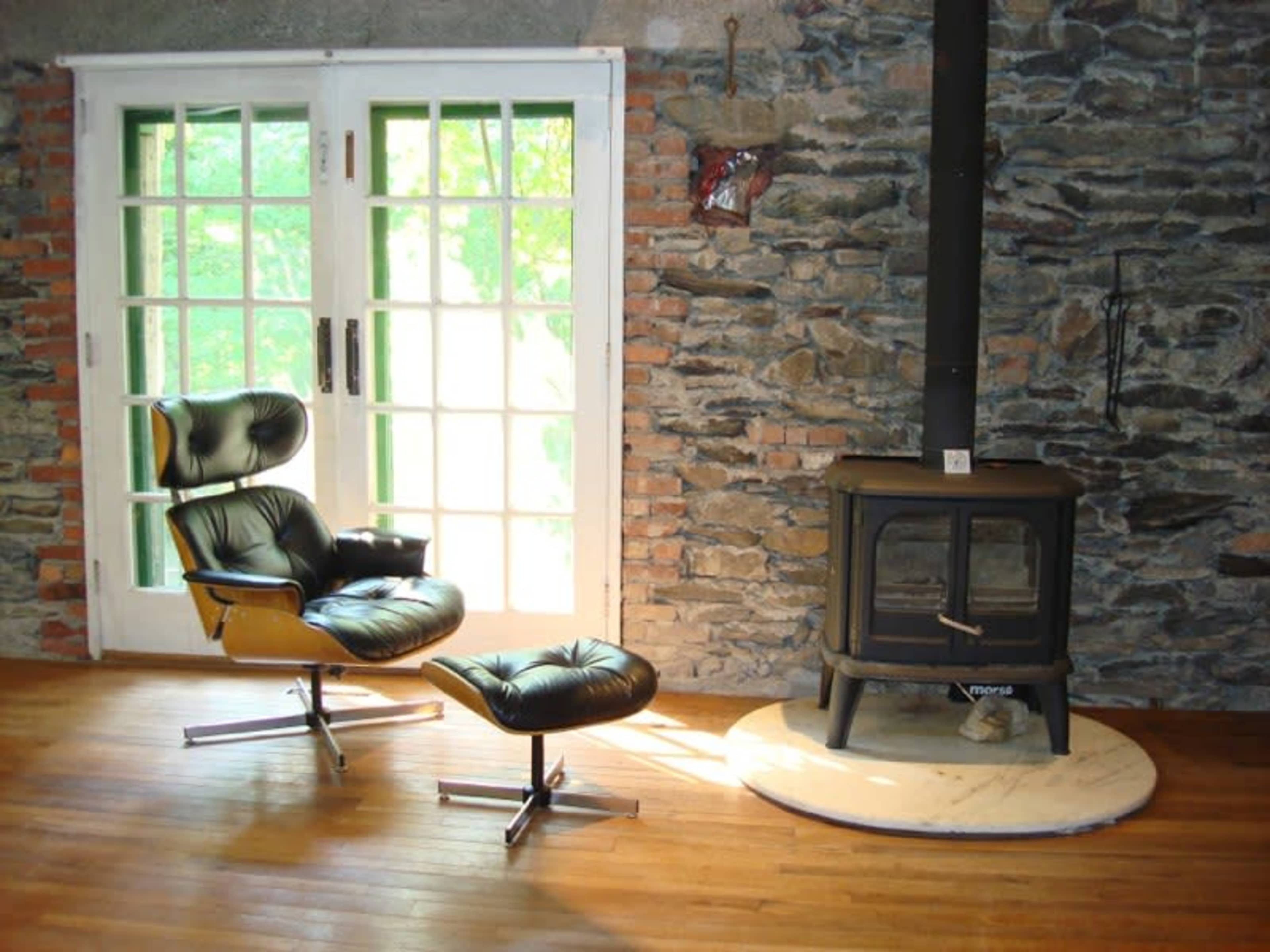 A black leather chair and ottoman sit beside a black metal wood stove on a marble pedestal, with large windows revealing a stone wall backdrop.