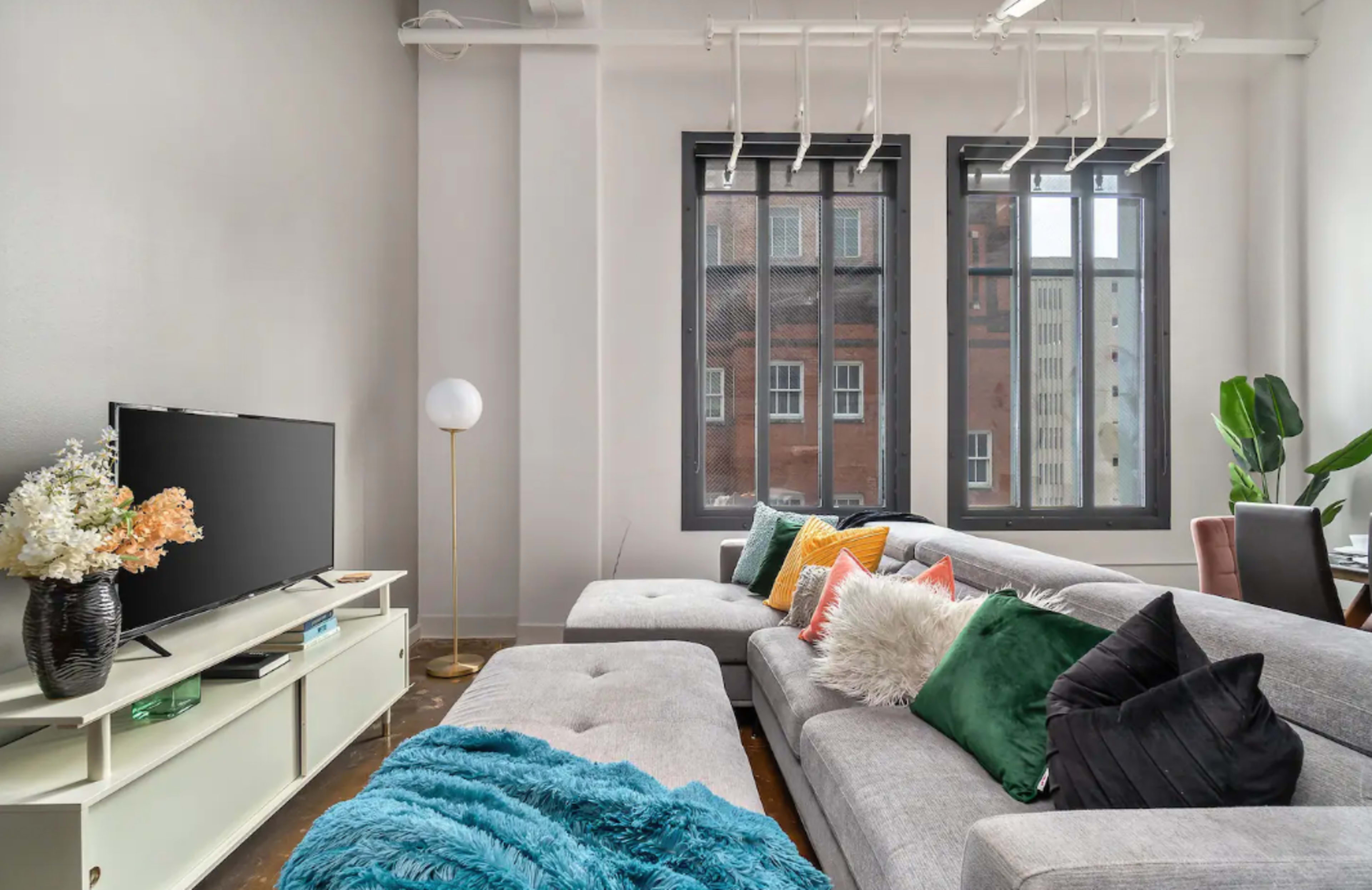 A modern living room featuring a sectional sofa with various pillows, a TV stand with a television, and large windows allowing natural light to fill the space.