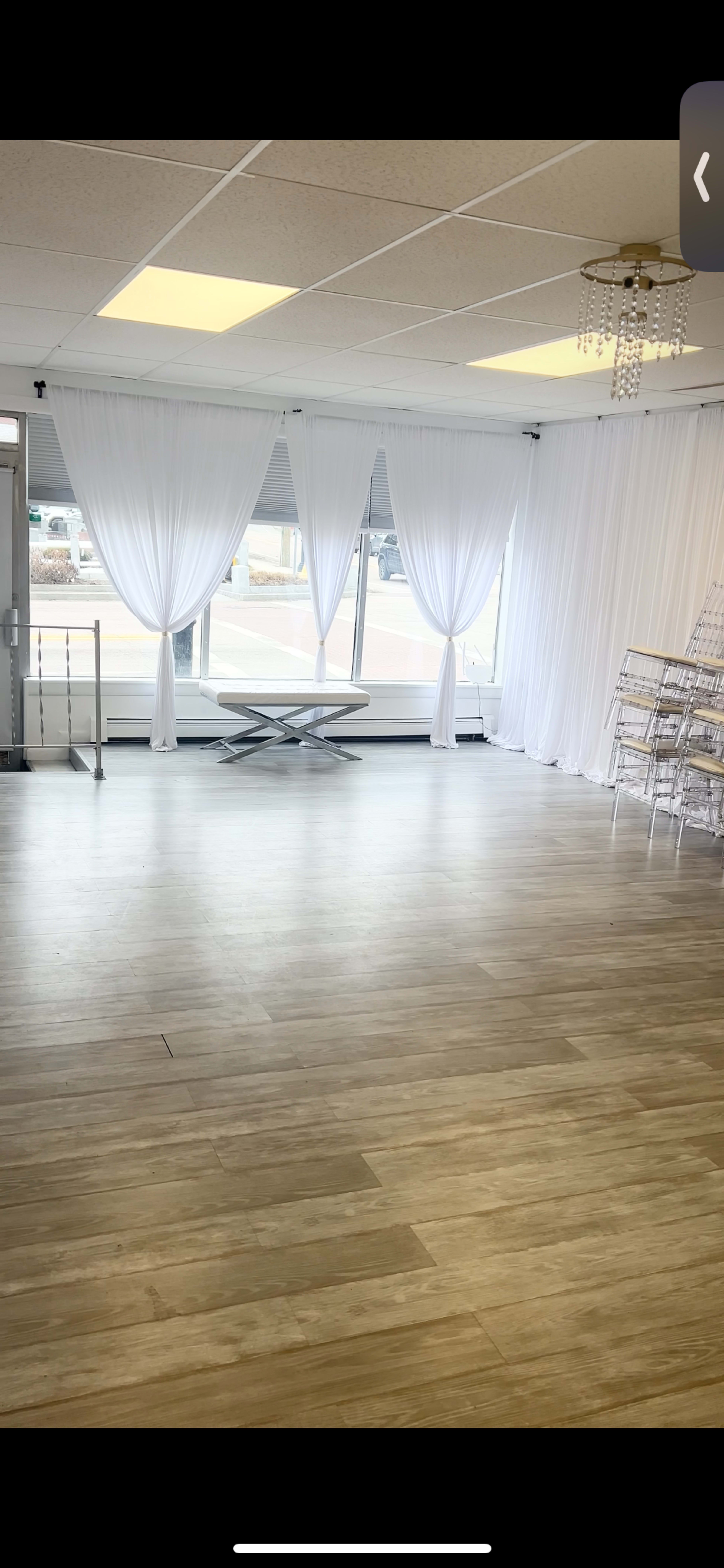 A spacious room features large windows with white drapes, a modern table in the center, and wooden flooring.