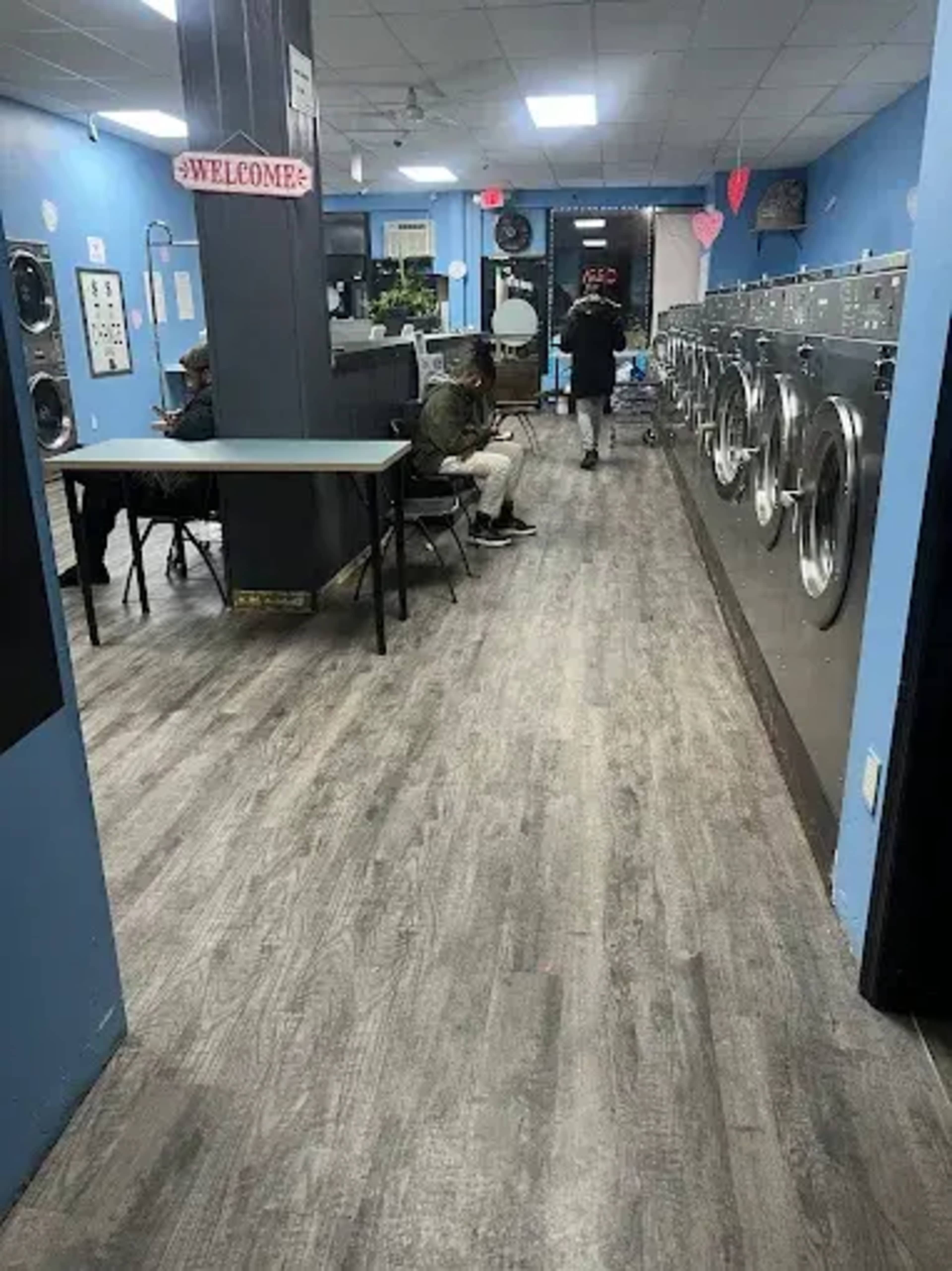 The image shows a laundromat with washing machines along one side and customers seated at tables on the opposite side.
