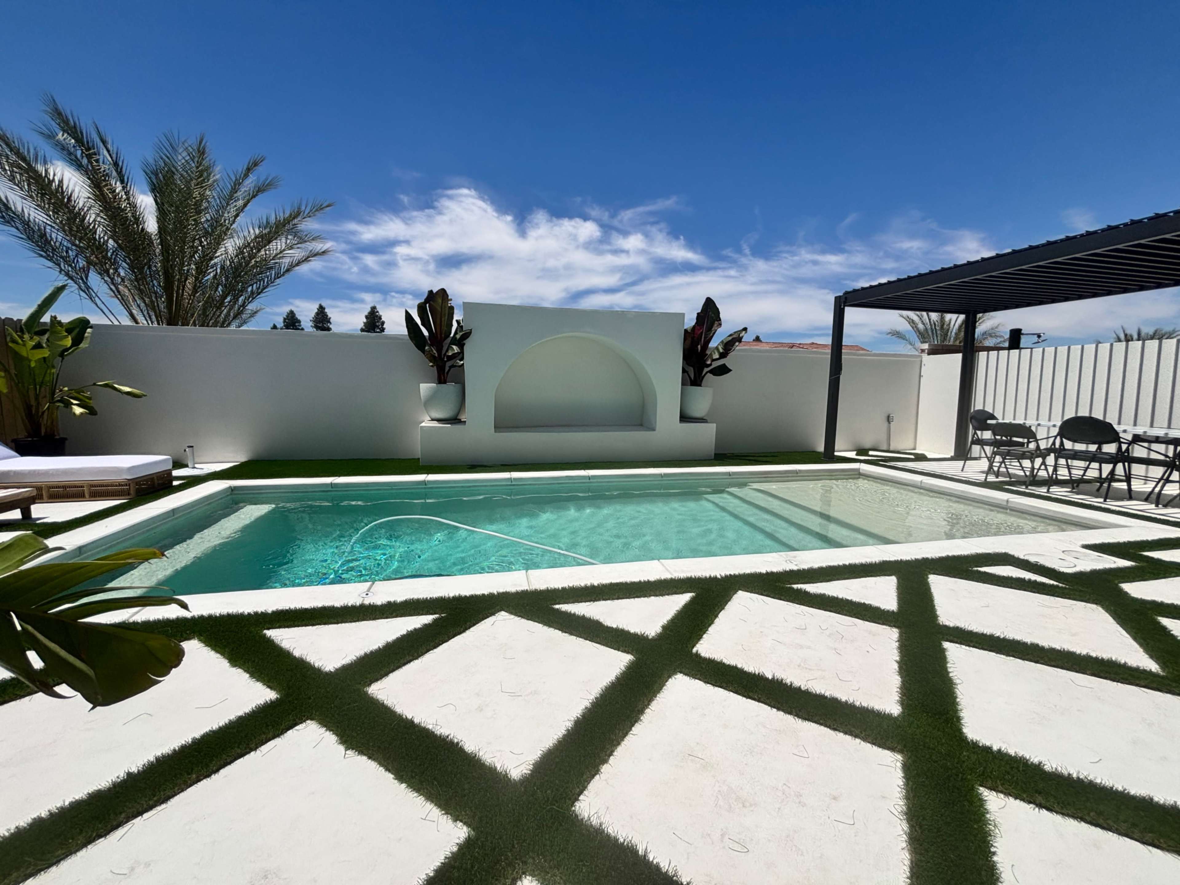 The image shows a modern swimming pool surrounded by greenery, with a seating area and a backdrop of palm trees against a clear blue sky.