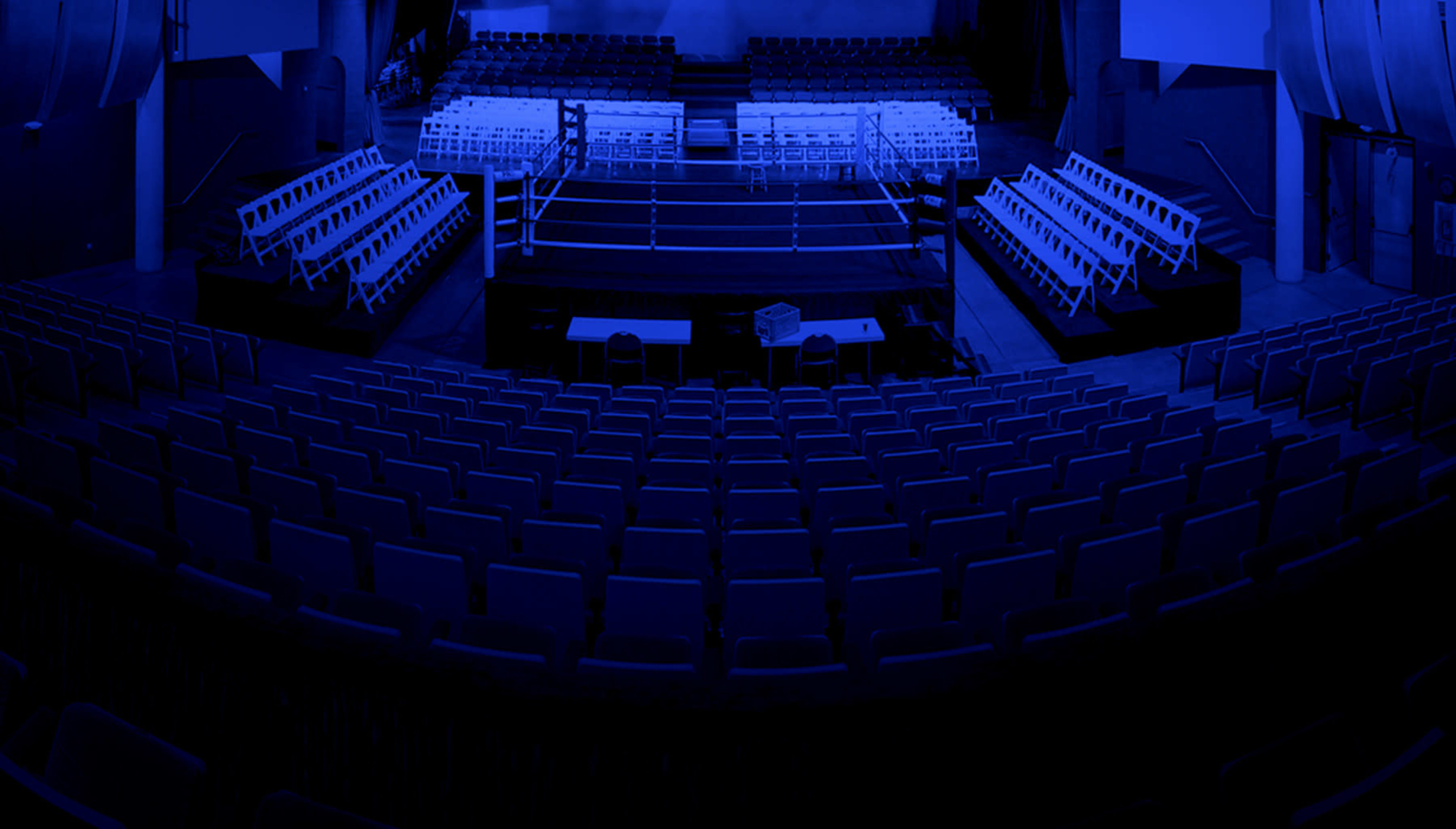 A boxing ring is set up in an empty arena, surrounded by rows of folding chairs under dim blue lighting.