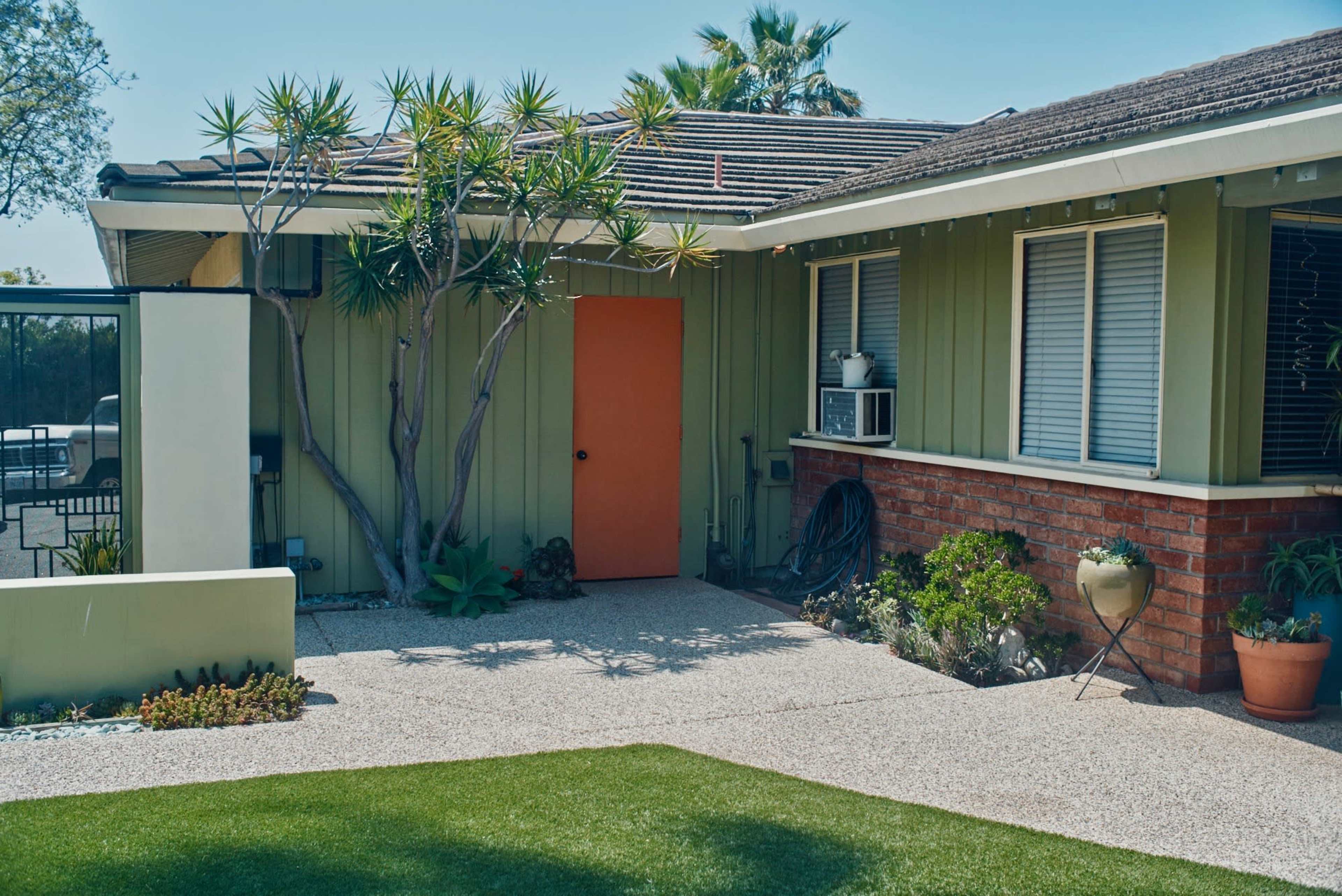 A house features an orange door, surrounded by green walls and landscaping, with a small garden and potted plants in the front yard.