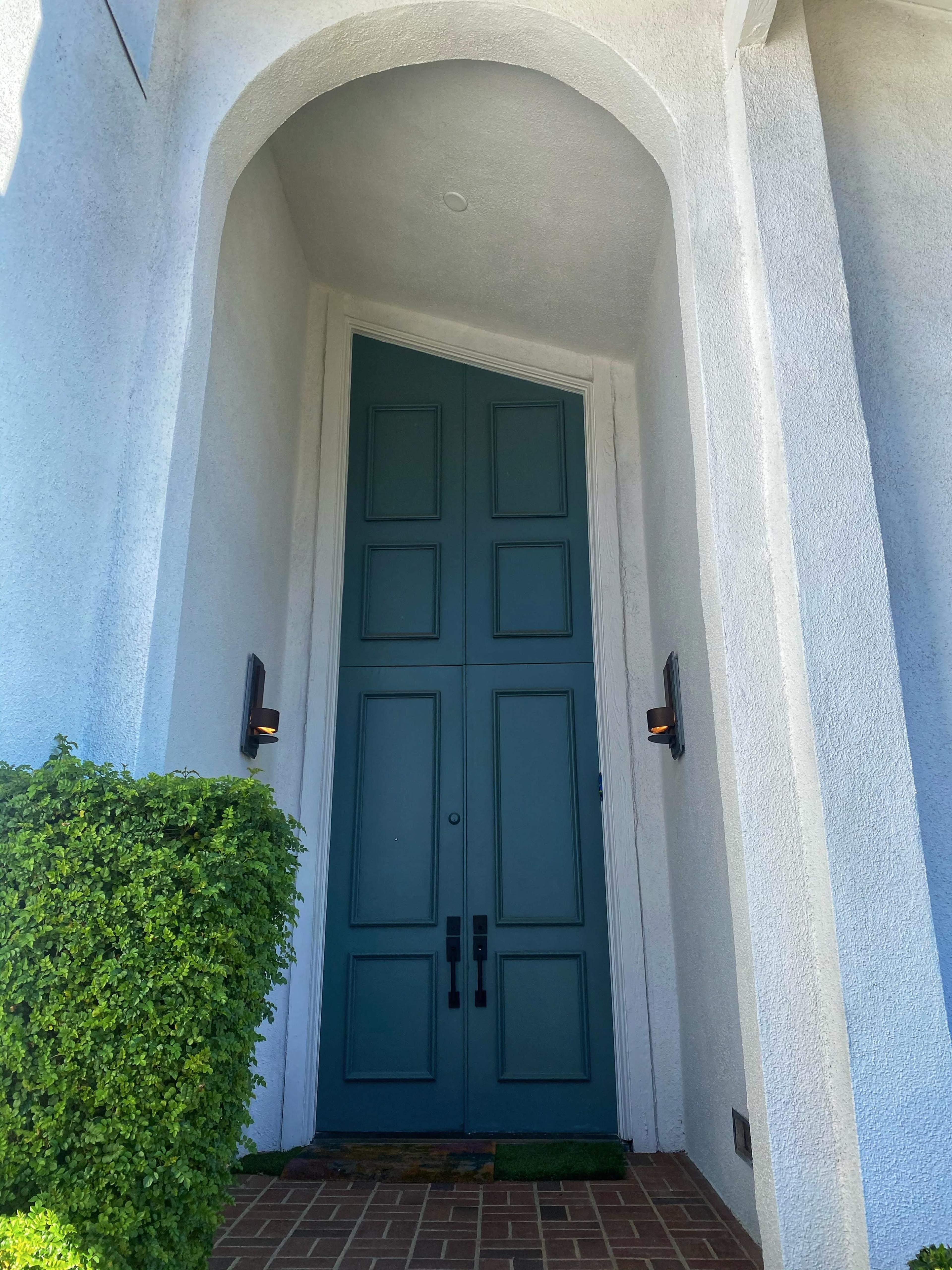 A tall, green double door is set within a white arched entryway, flanked by two wall sconces and bordered by neatly trimmed greenery.