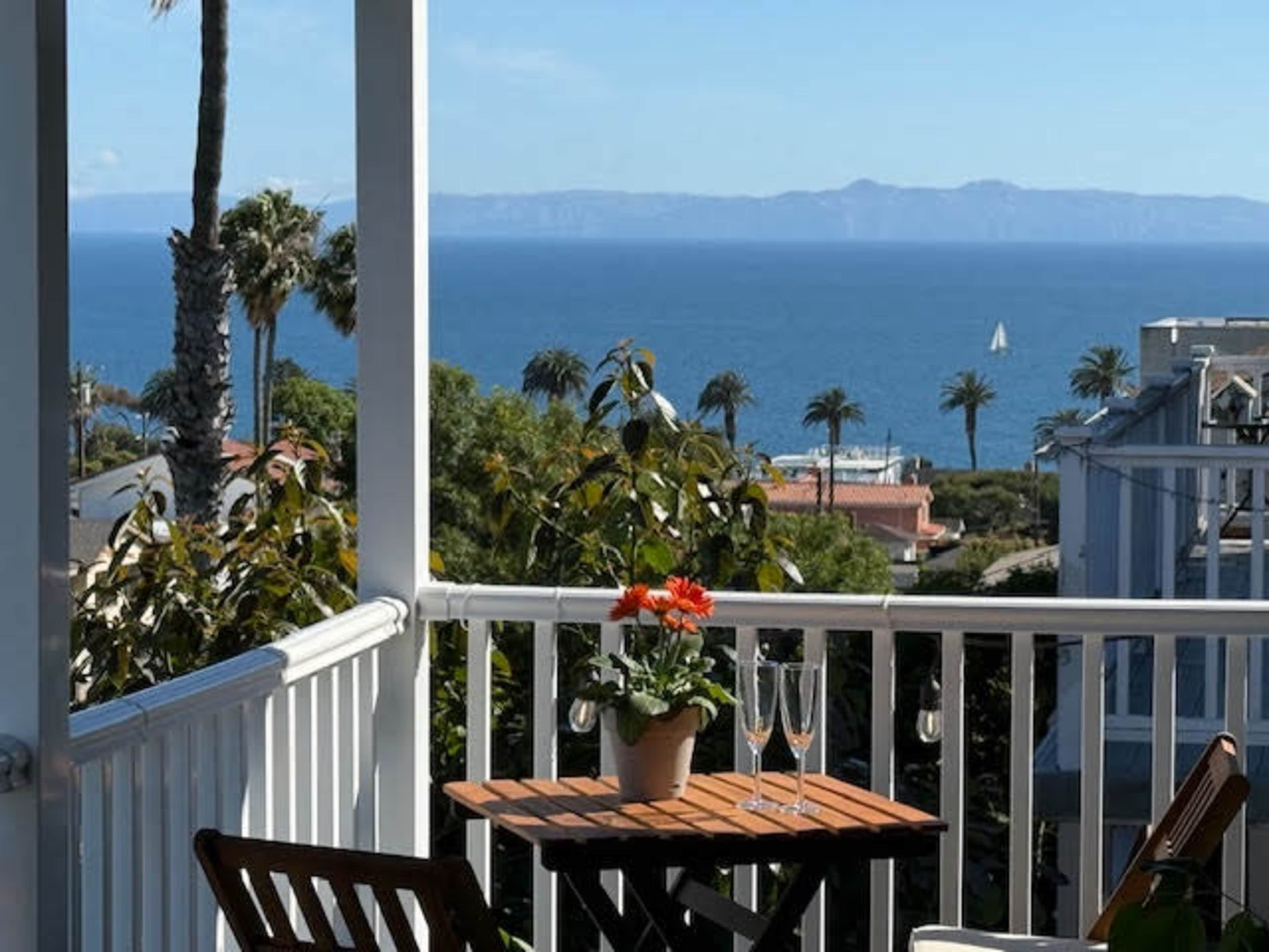 The image shows a balcony with a table set for two, featuring a potted flower and two glasses, overlooking a calm sea and distant mountains.