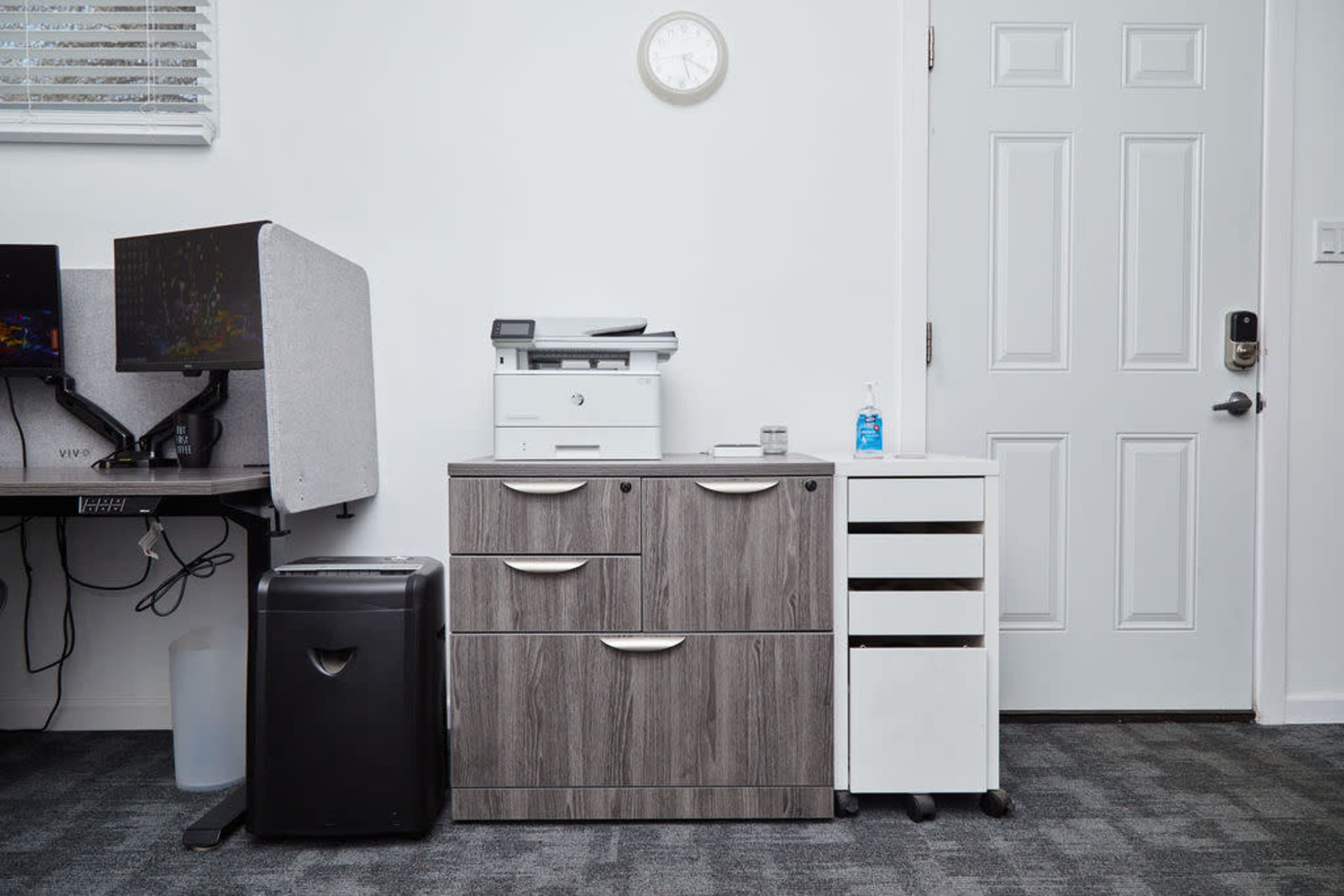 An office corner featuring a gray filing cabinet, a white printer, a small mobile filing unit, a black trash can, and a white door.