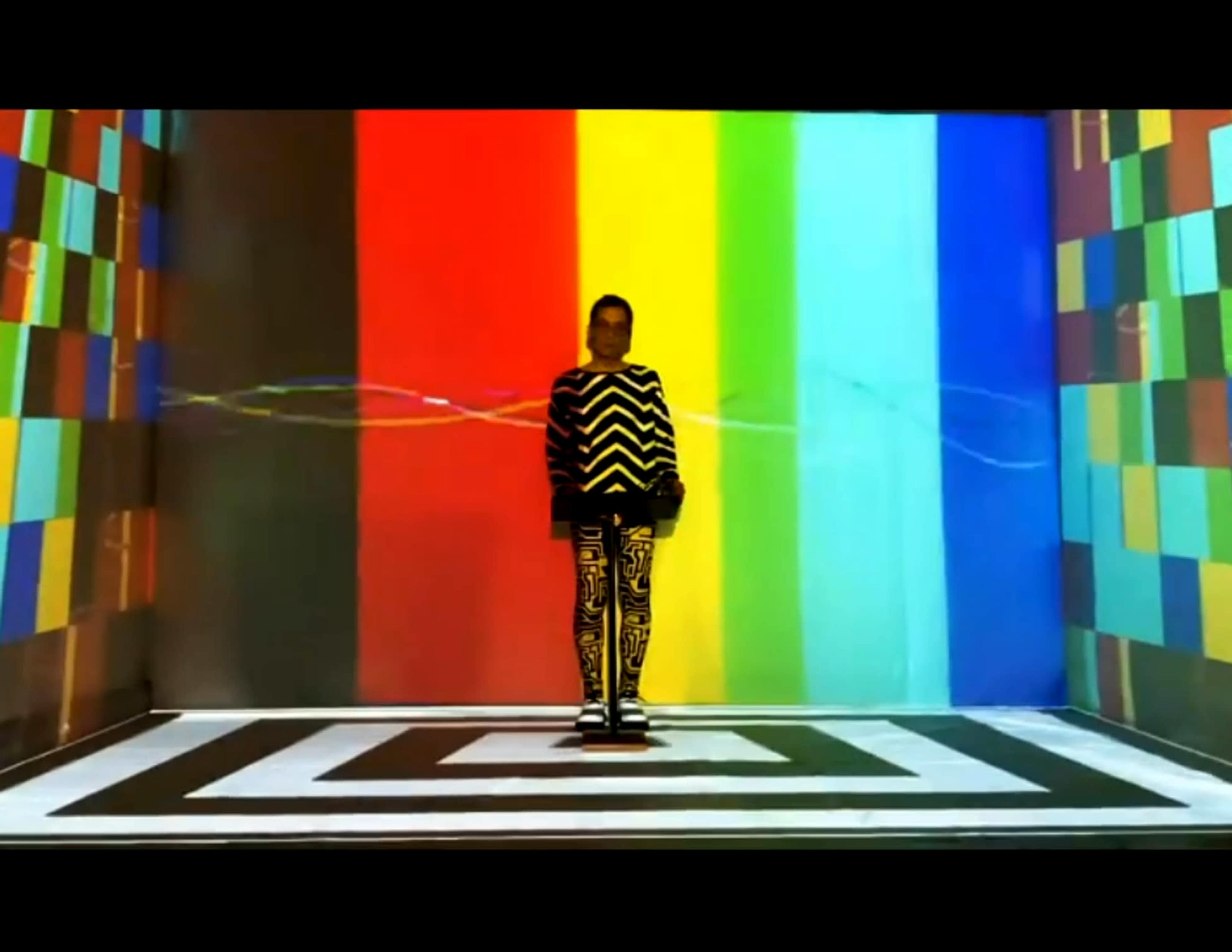 A figure stands on a geometric patterned floor in front of brightly colored vertical stripes and a colorful background.