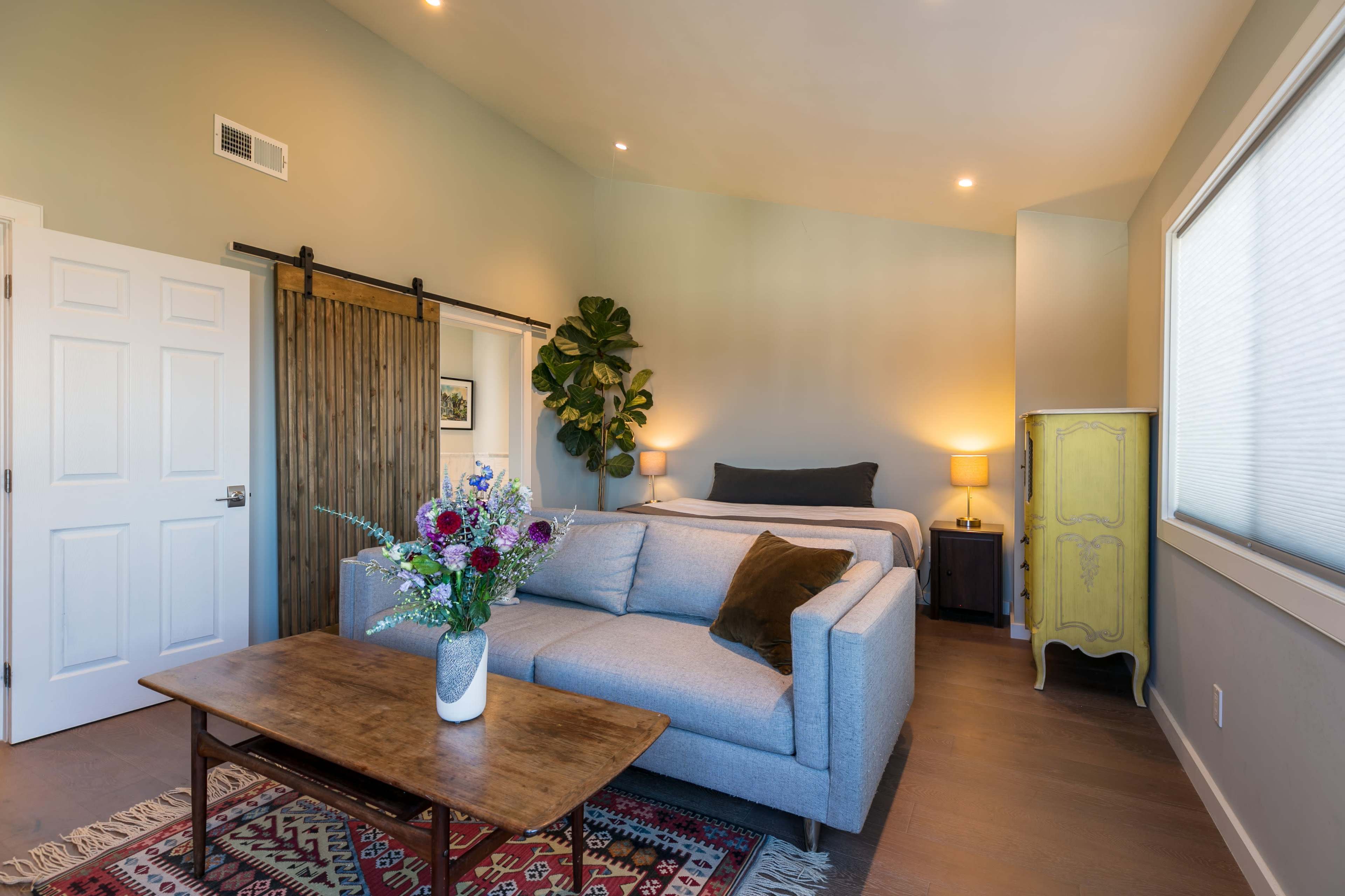 A cozy bedroom features a gray sofa, a wooden coffee table, and a bed with two bedside lamps, set against a backdrop of natural light and decorative plants.