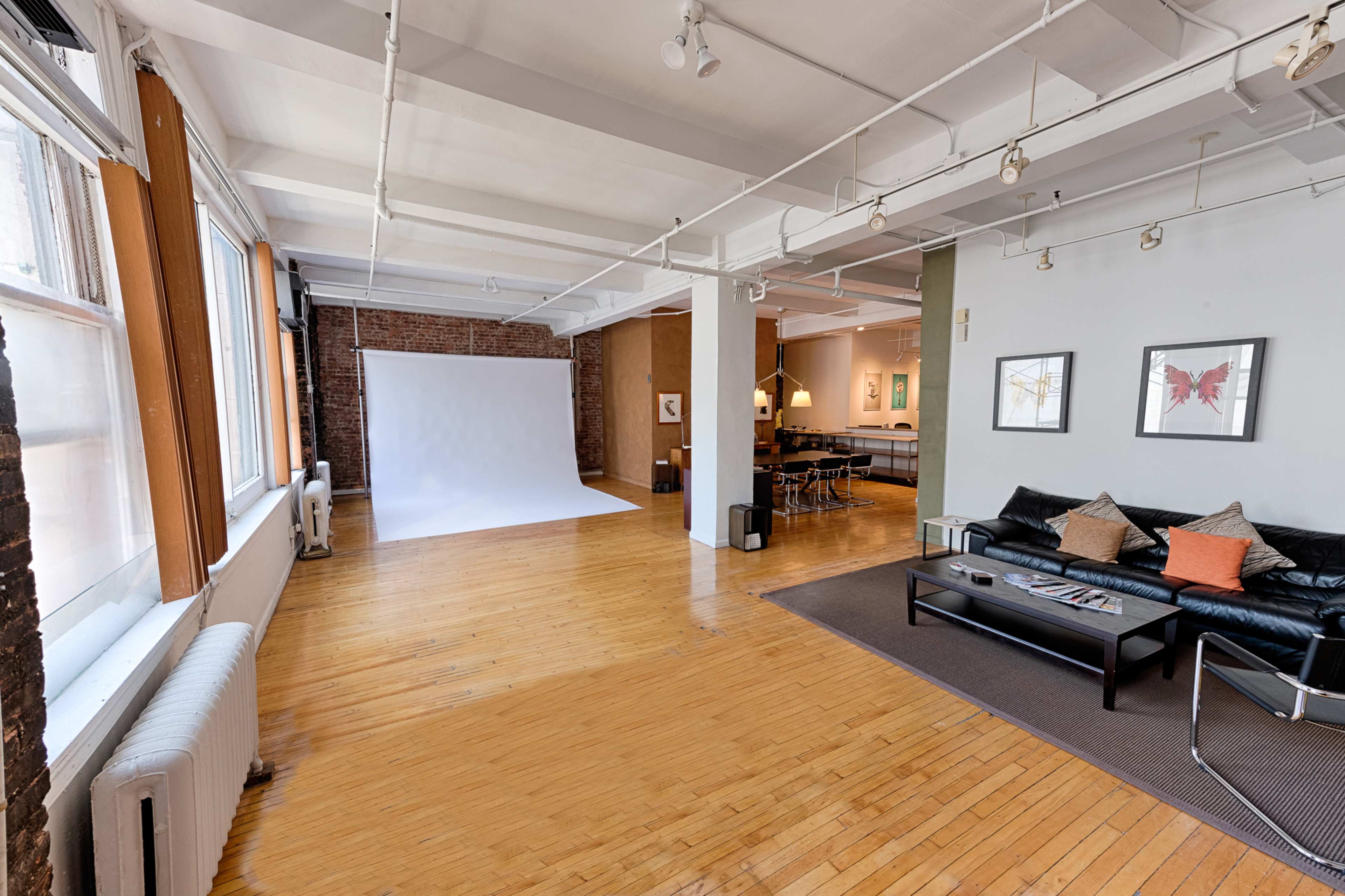 A spacious room with hardwood floors, featuring a white backdrop, a black leather couch, and modern furniture arranged for a cozy, open layout.