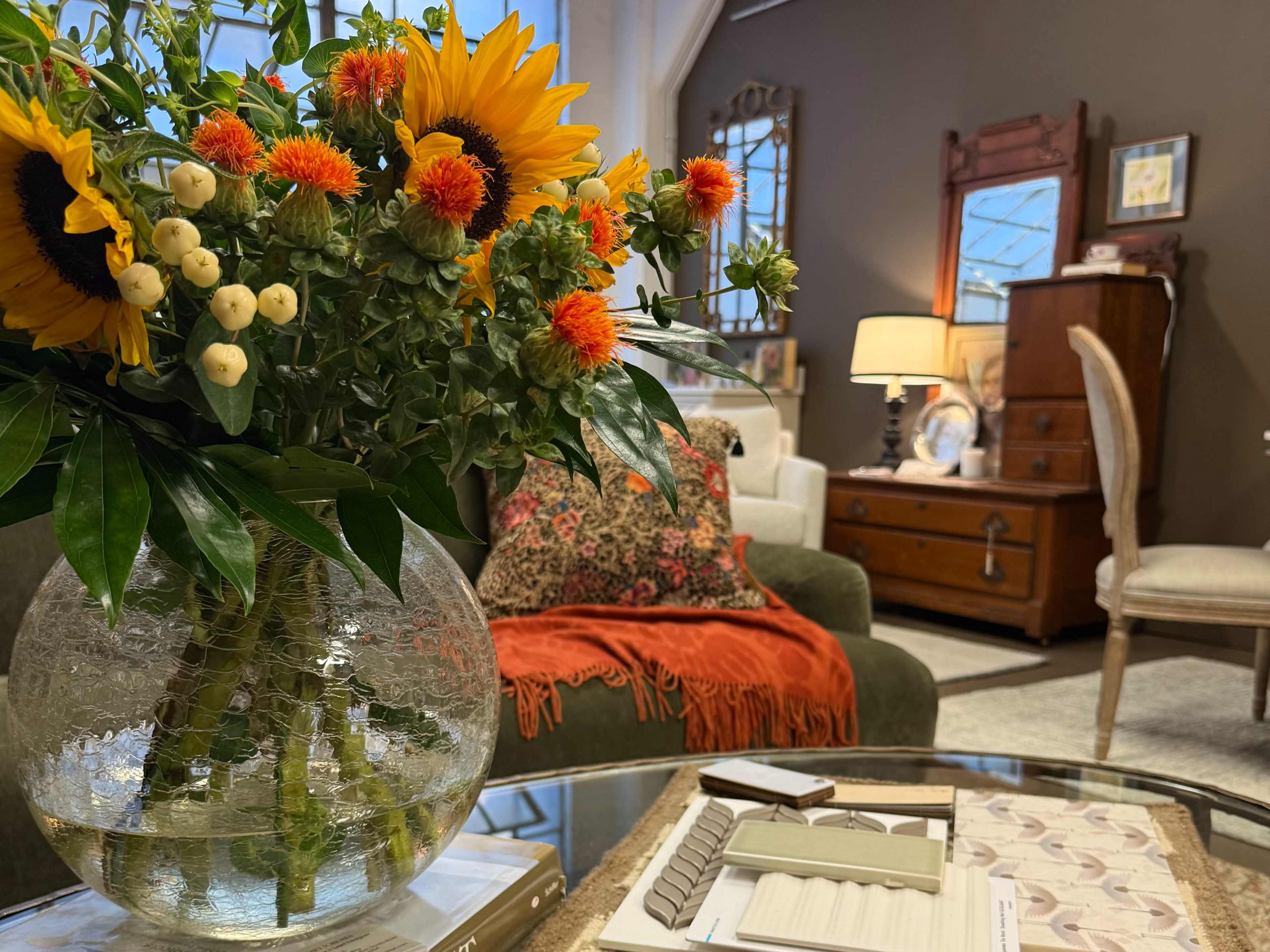 A vase of vibrant flowers sits on a glass table in a cozy living room with a green couch and an antique dresser.
