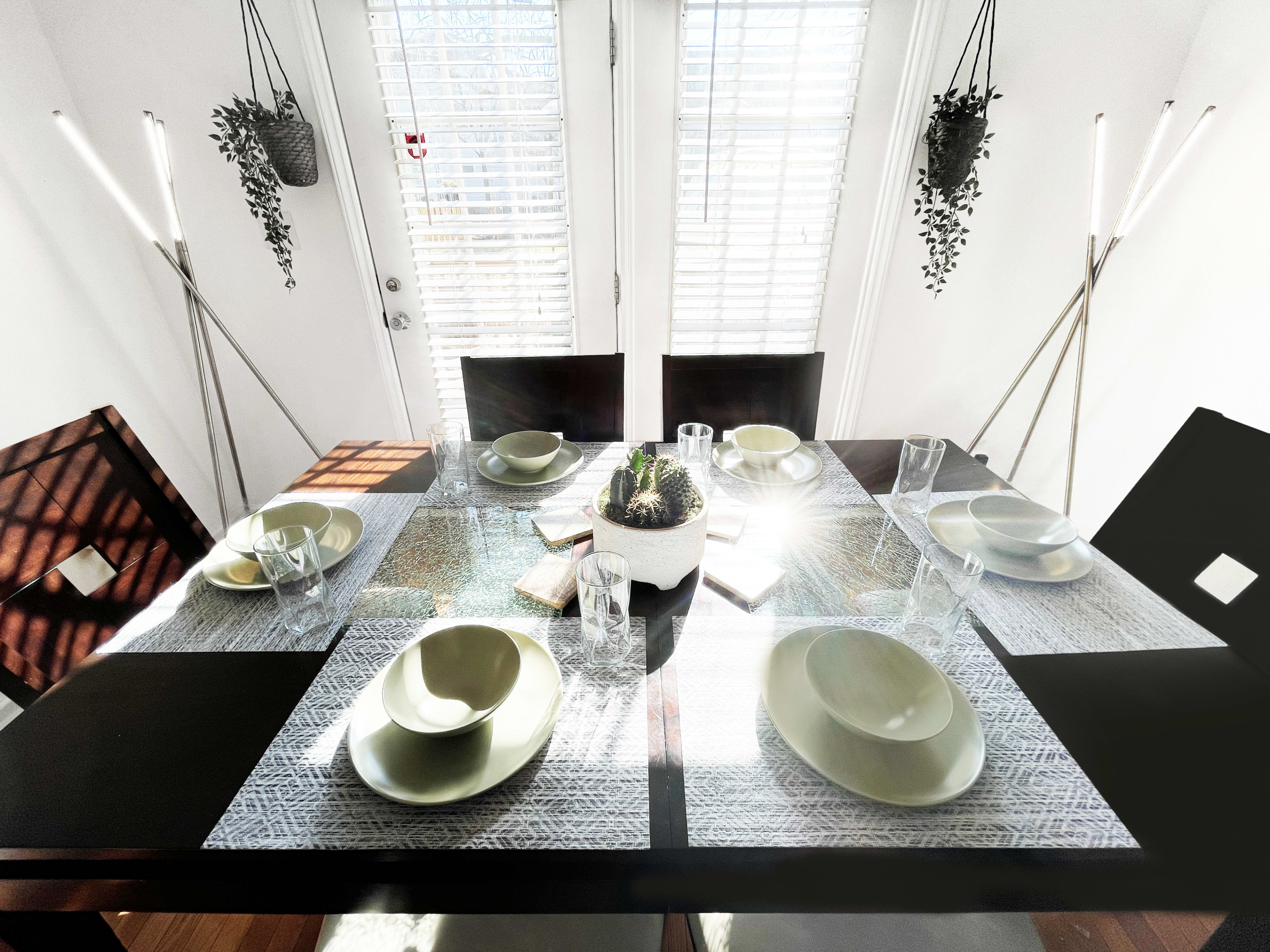 A dining table is set with green plates and glasses, surrounded by bright natural light filtering through window shutters.