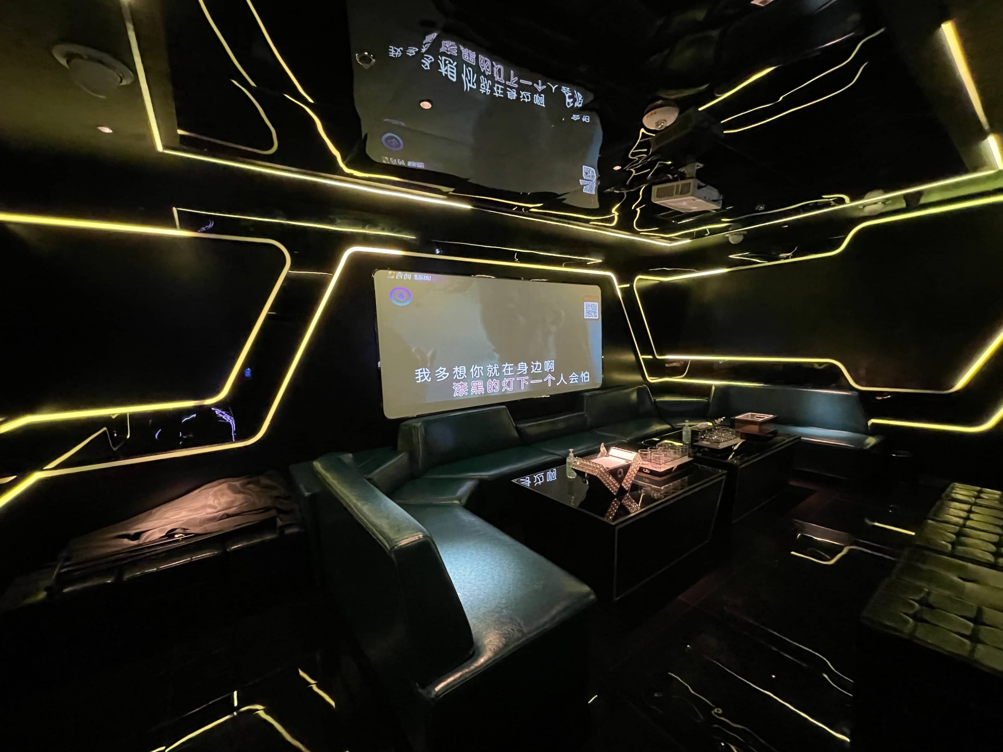 A modern karaoke room featuring black walls, neon lighting, a large screen displaying text, and a sleek sofa arrangement around a central coffee table.