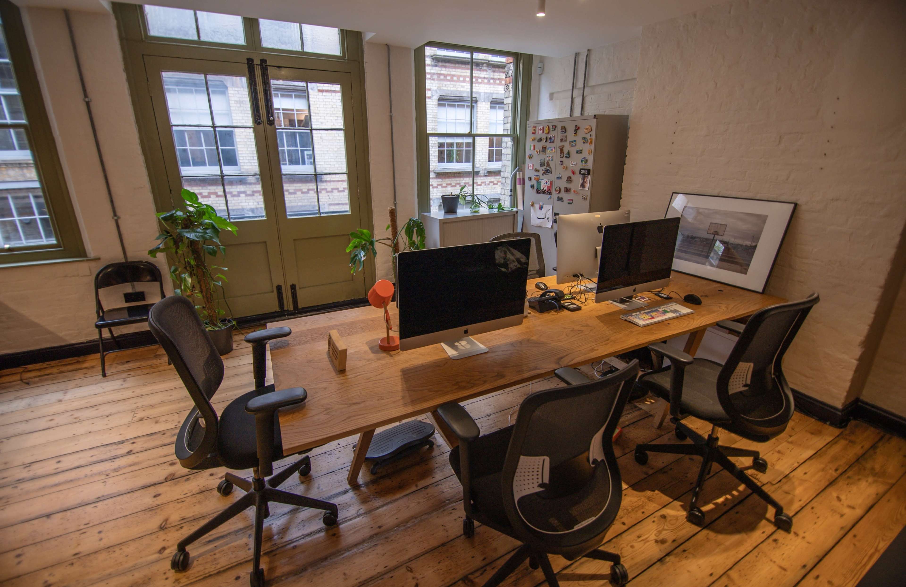 A modern office space features a large wooden desk with multiple computer monitors, surrounded by black office chairs, and illuminated by natural light from large windows.