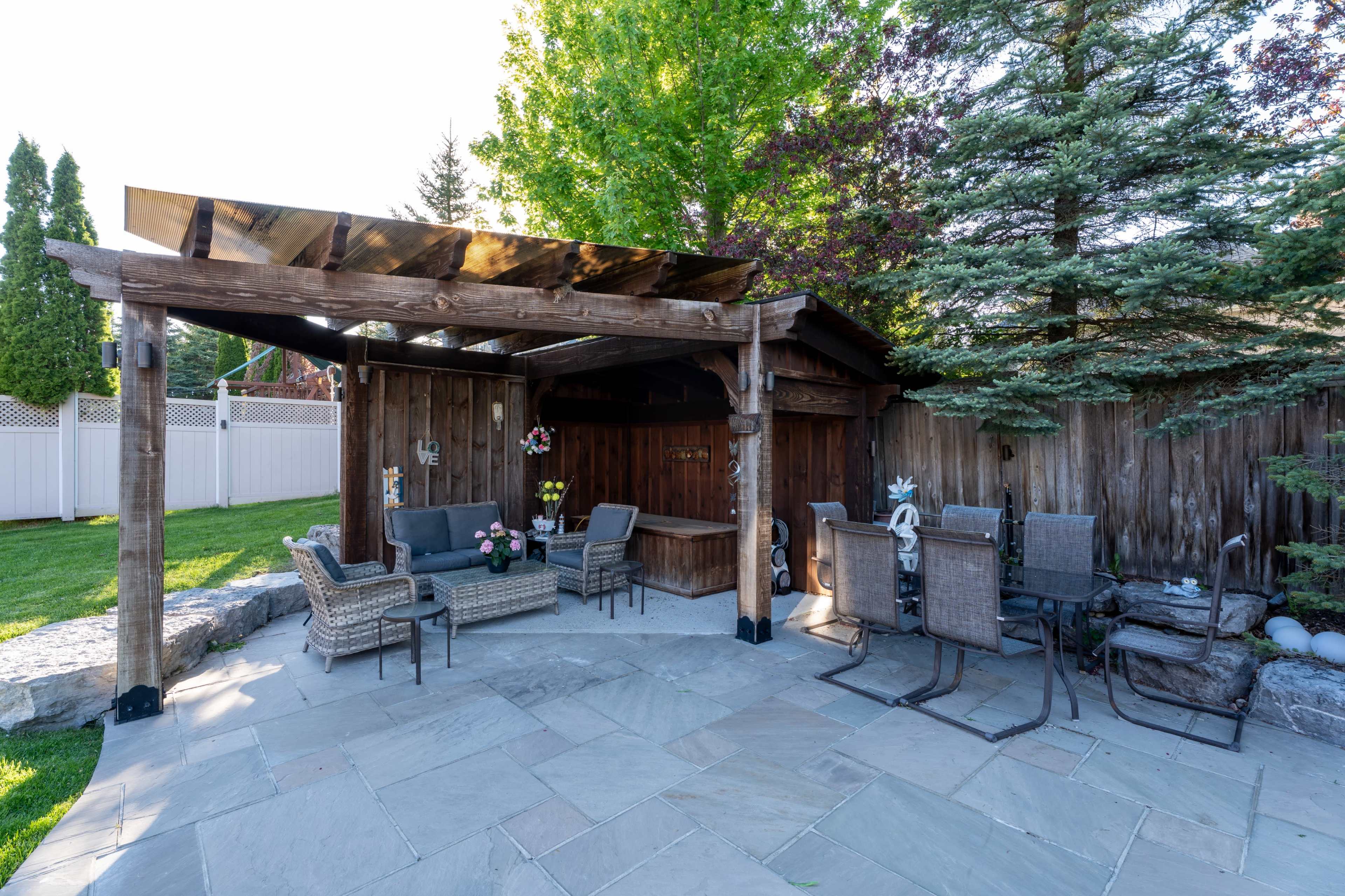 The scene features a landscaped backyard with a wooden pergola, comfortable seating, and a stone patio surrounded by trees and a fence.