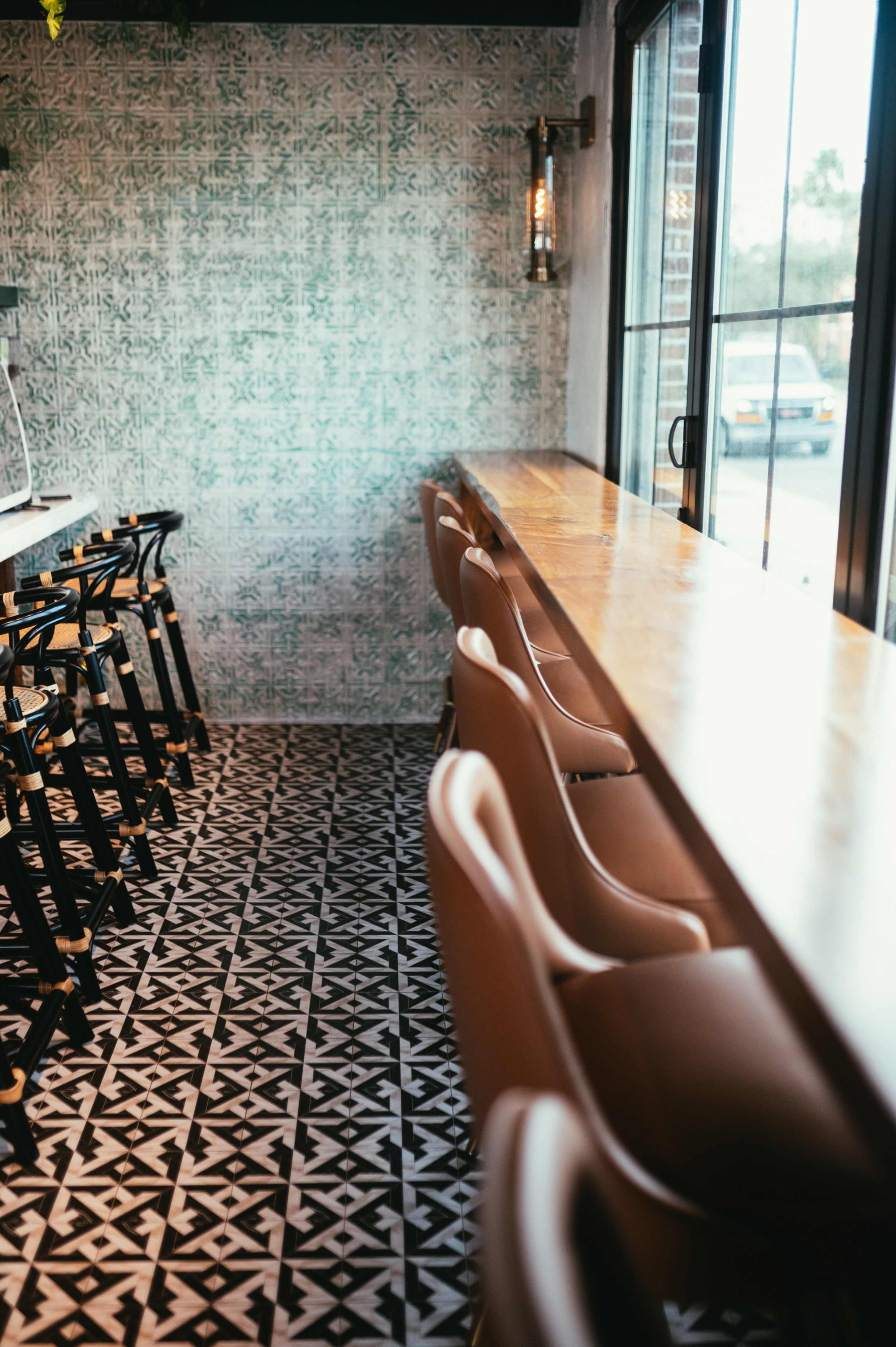 The image shows a modern bar area with a long wooden counter and brown upholstered stools, set against a patterned tile floor and a decorative wall.