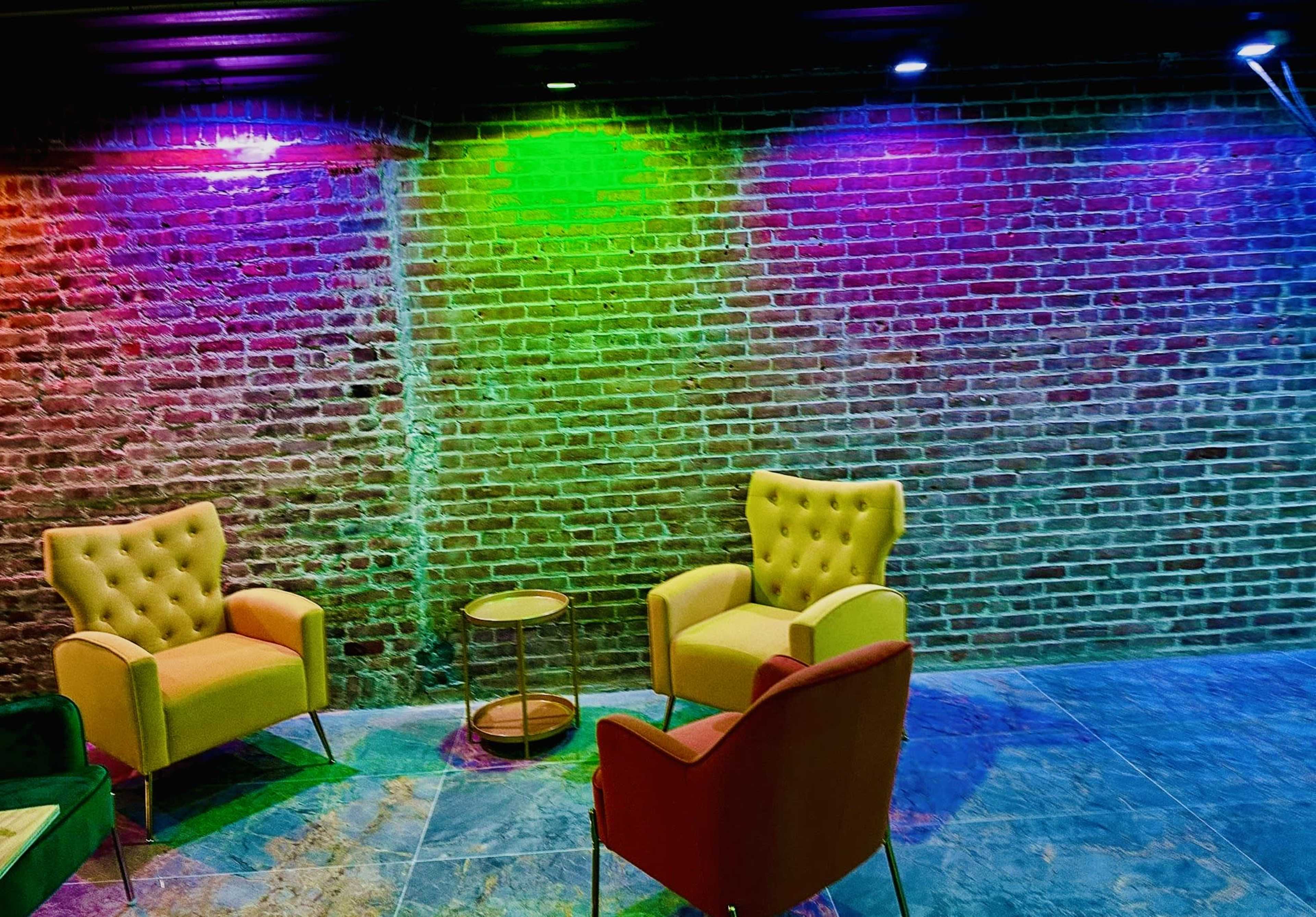 Two upholstered chairs in yellow and red, positioned near a small round table against a backdrop of a brick wall illuminated with colorful lights.