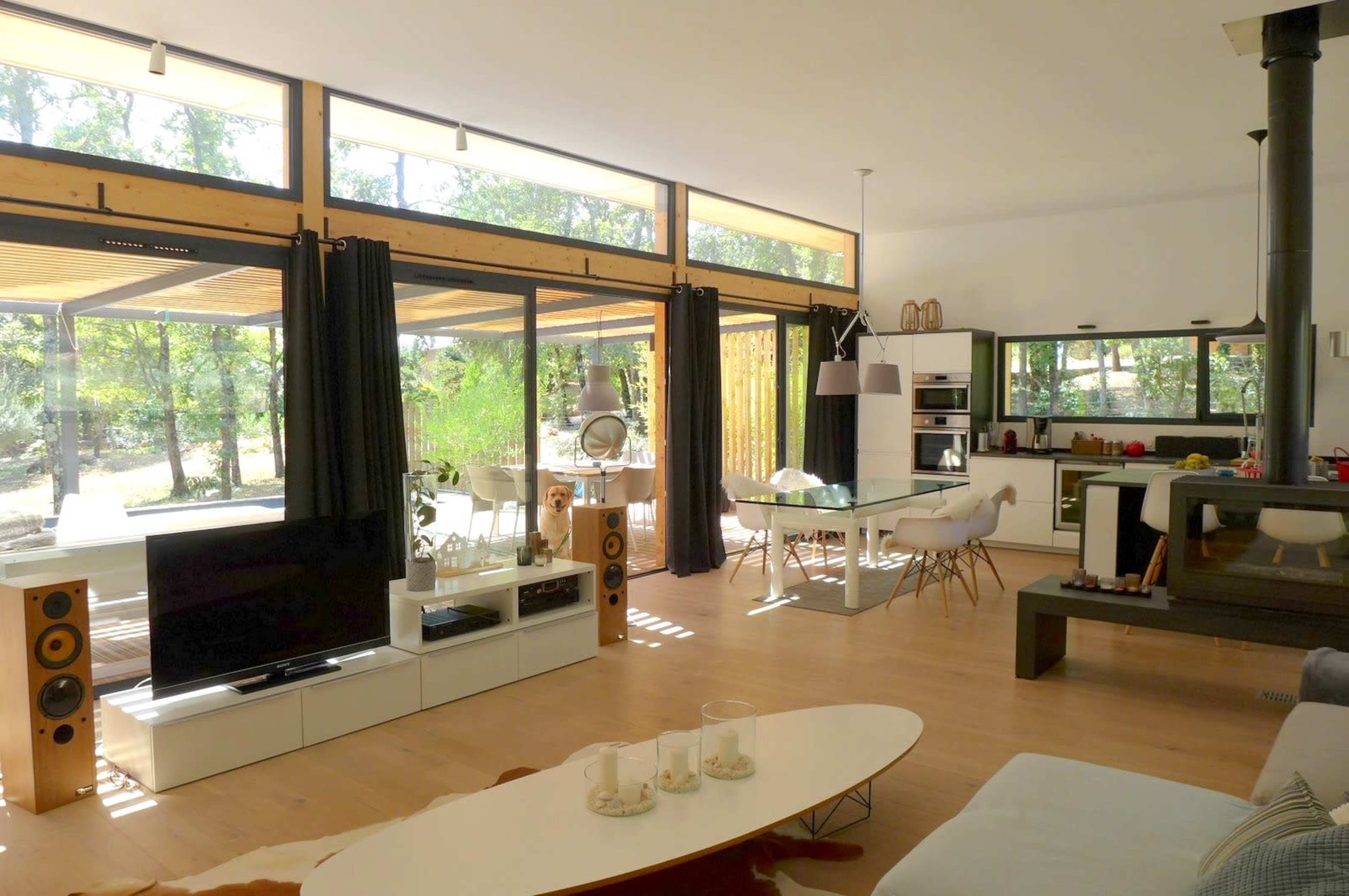 A modern, open-concept living area with large windows, light-colored walls, a dining table, and a sleek kitchen.