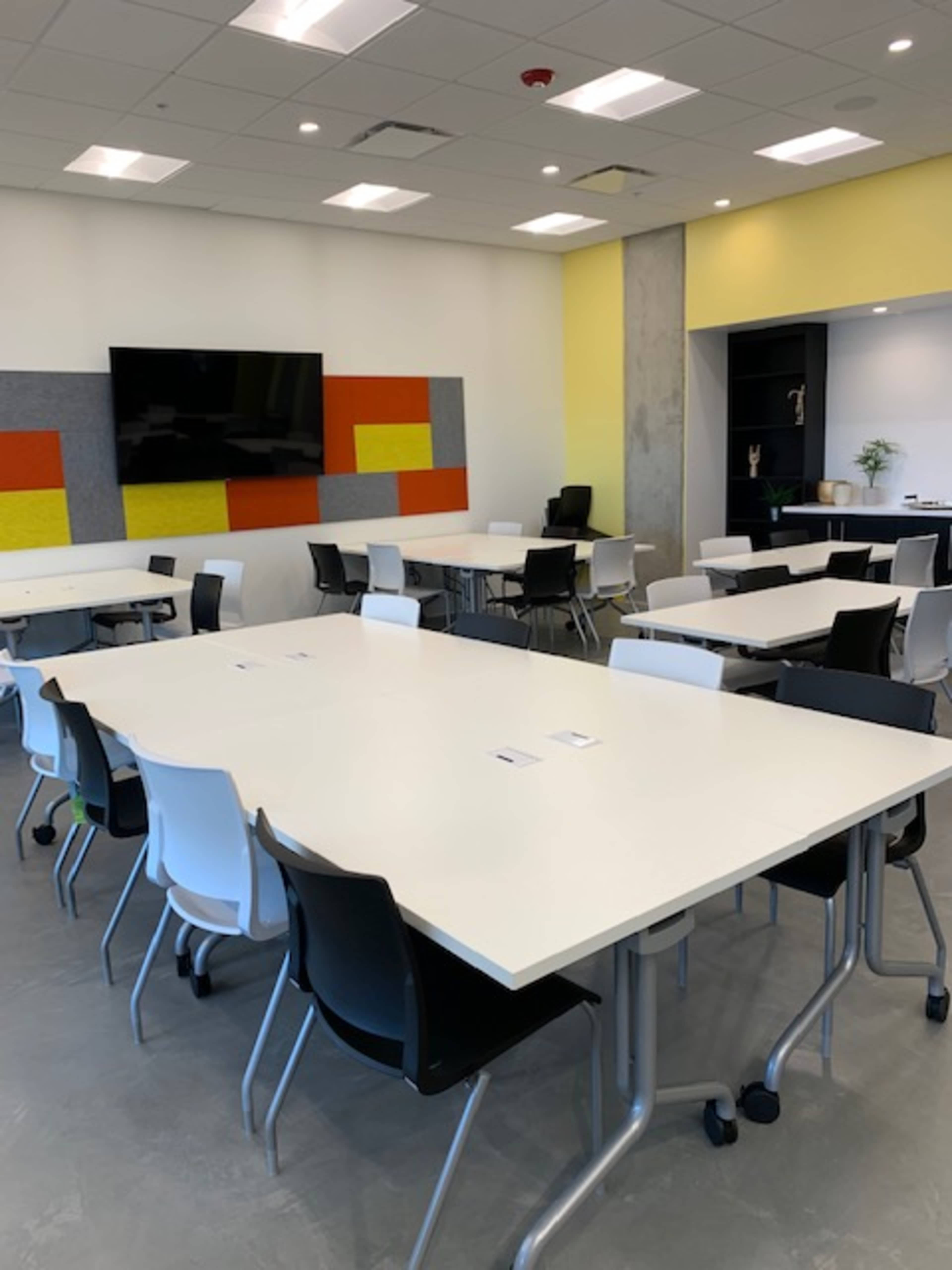 A modern conference room is arranged with multiple white tables and chairs, featuring a large television screen and colorful wall panels.