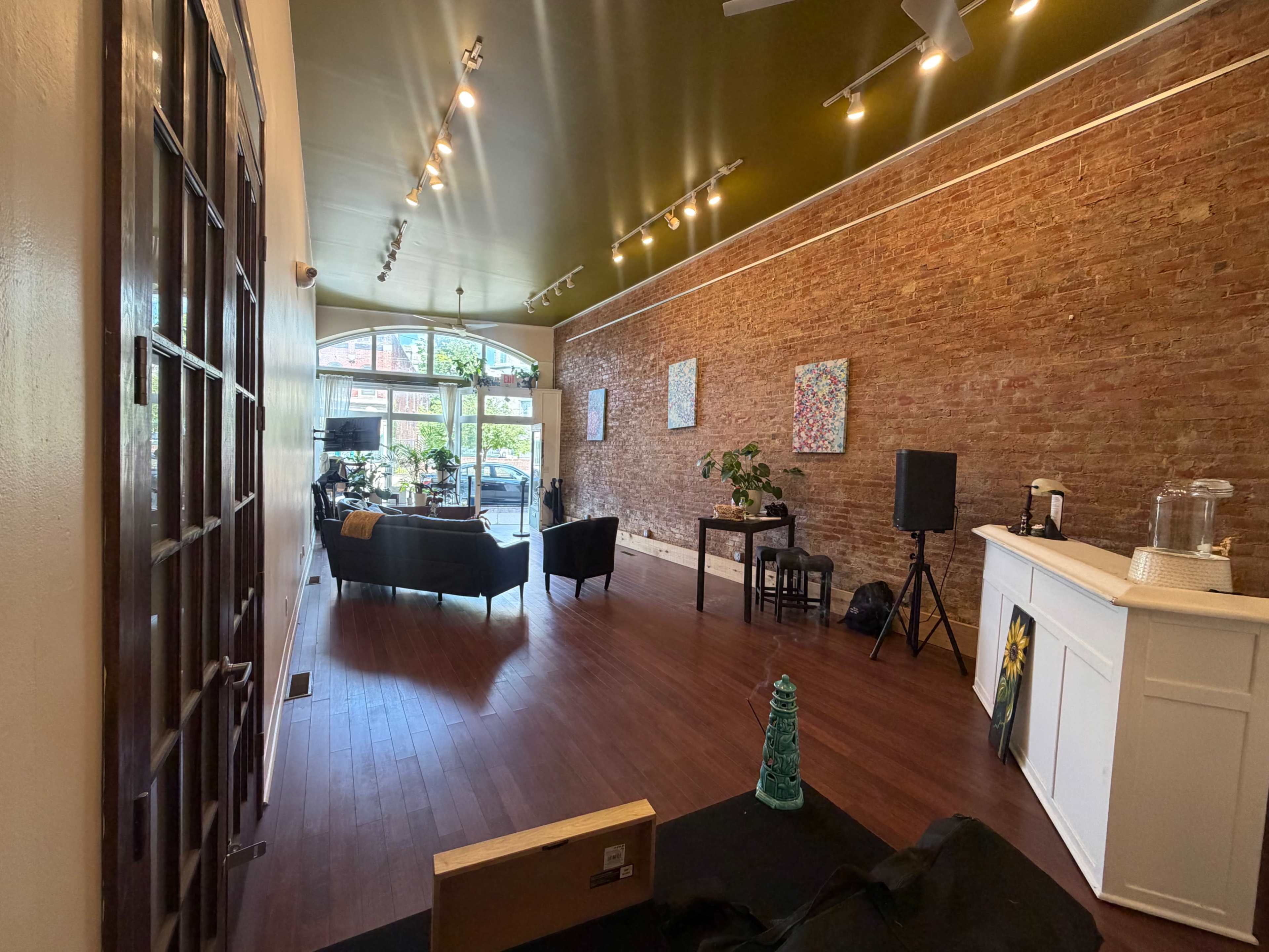 The image shows a spacious interior of a brick-walled room with hardwood floors, featuring seating areas, plants, and a service counter.