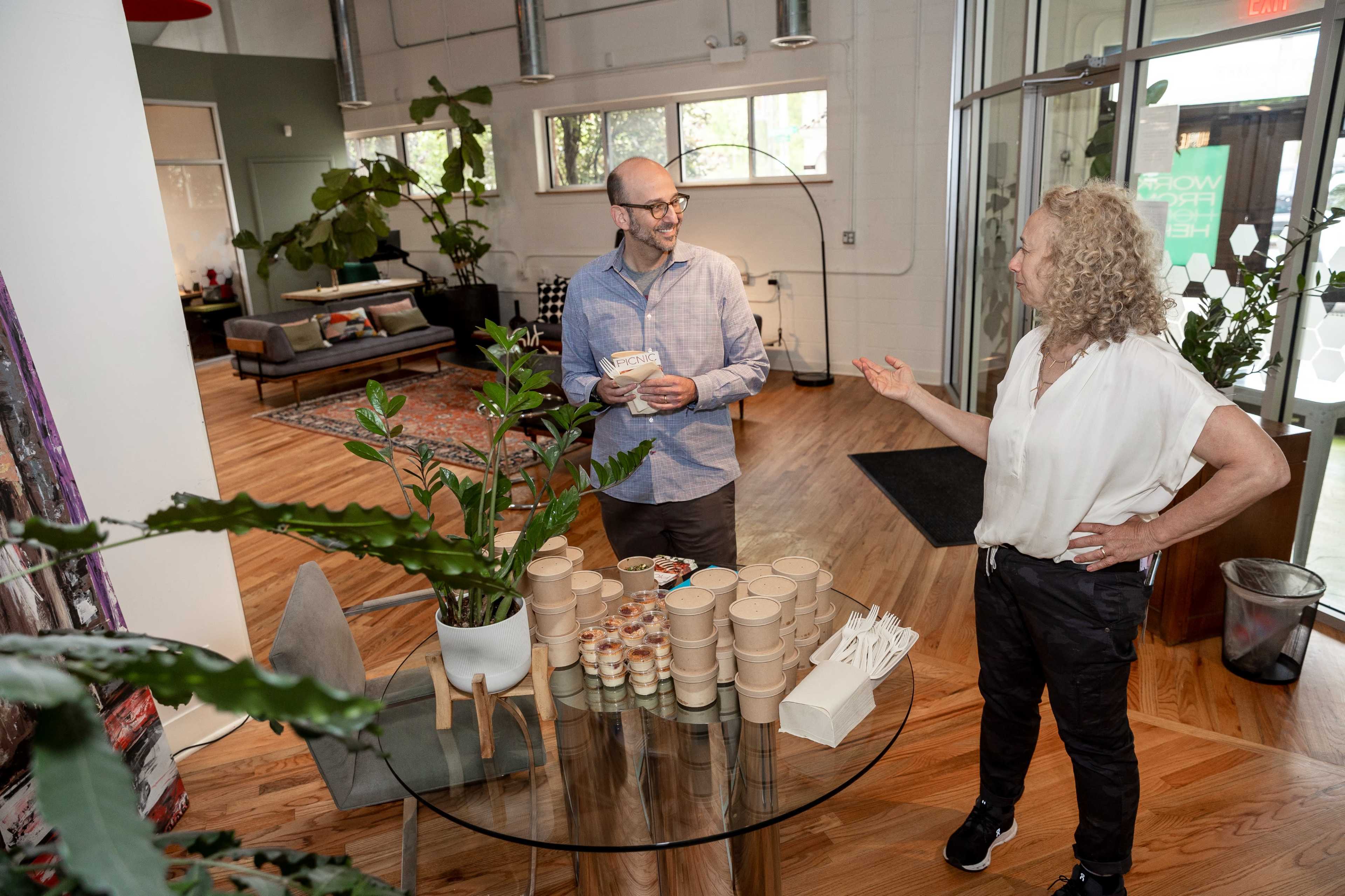 Two people are talking near a glass table with containers and utensils in a modern, plant-filled indoor space.