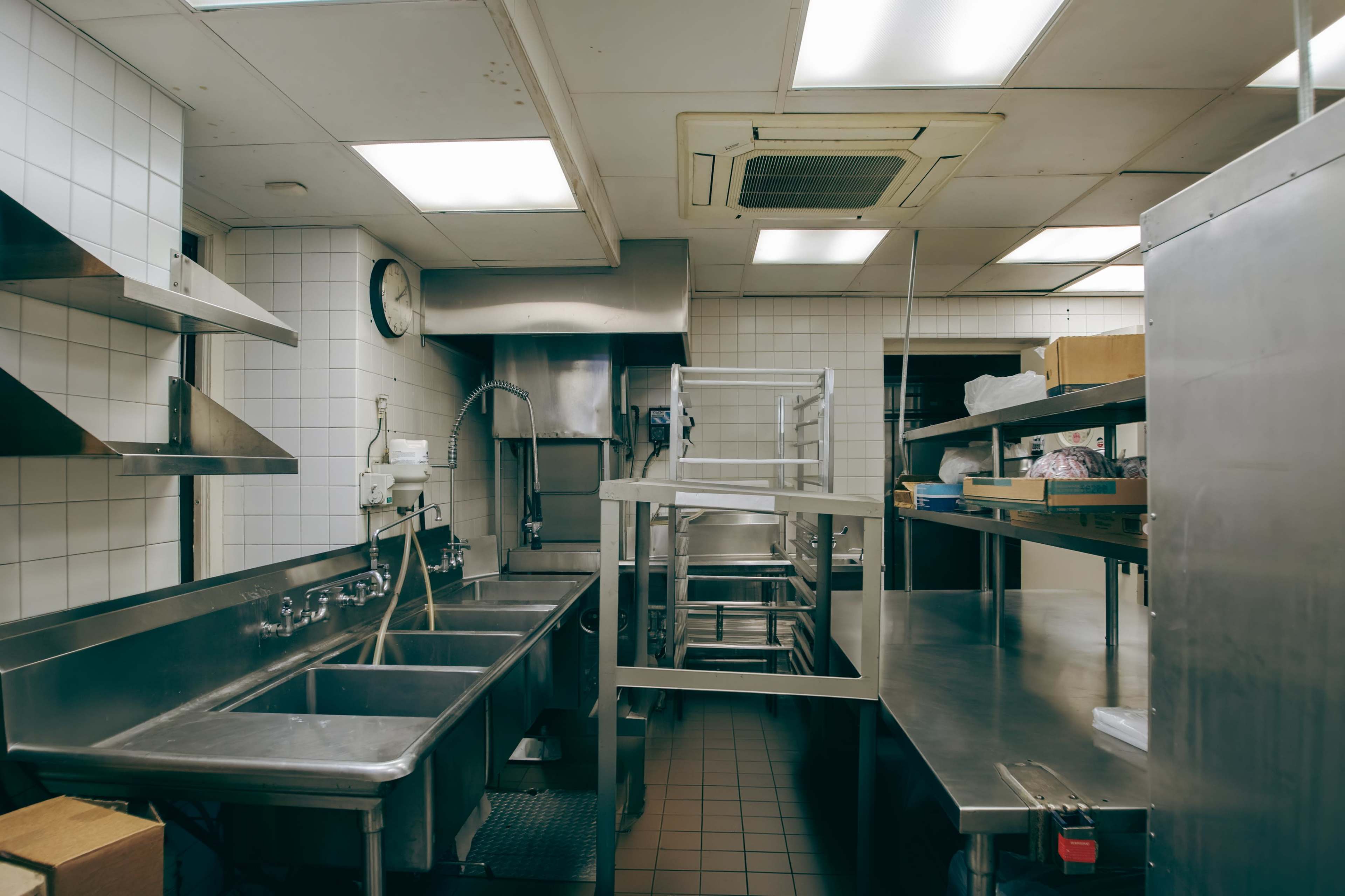 A commercial kitchen with stainless steel sinks, shelves, and cooking equipment against tiled walls and fluorescent lighting.