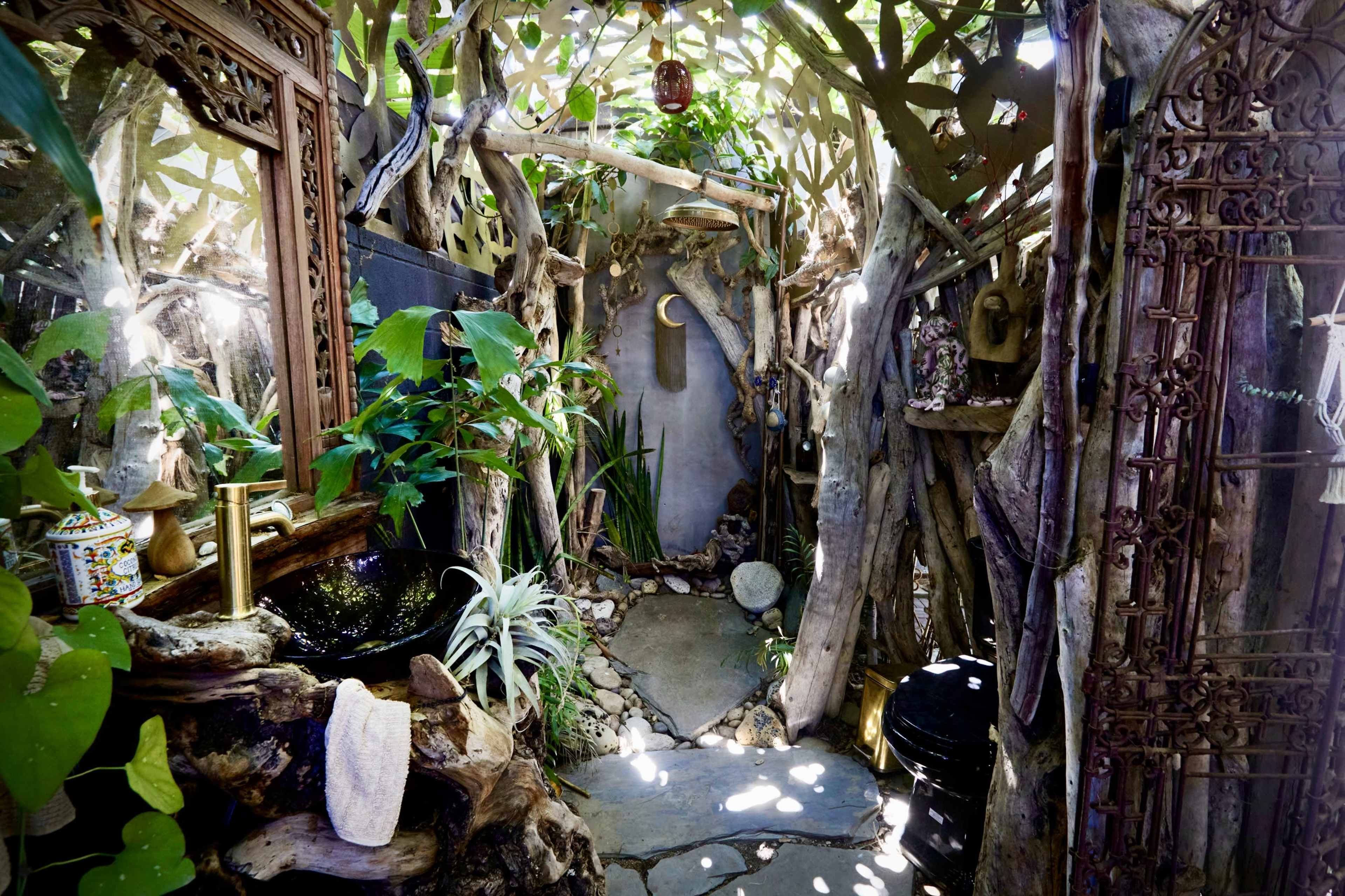 The image depicts a rustic outdoor shower area surrounded by natural wood structures, plants, and stone pathways.