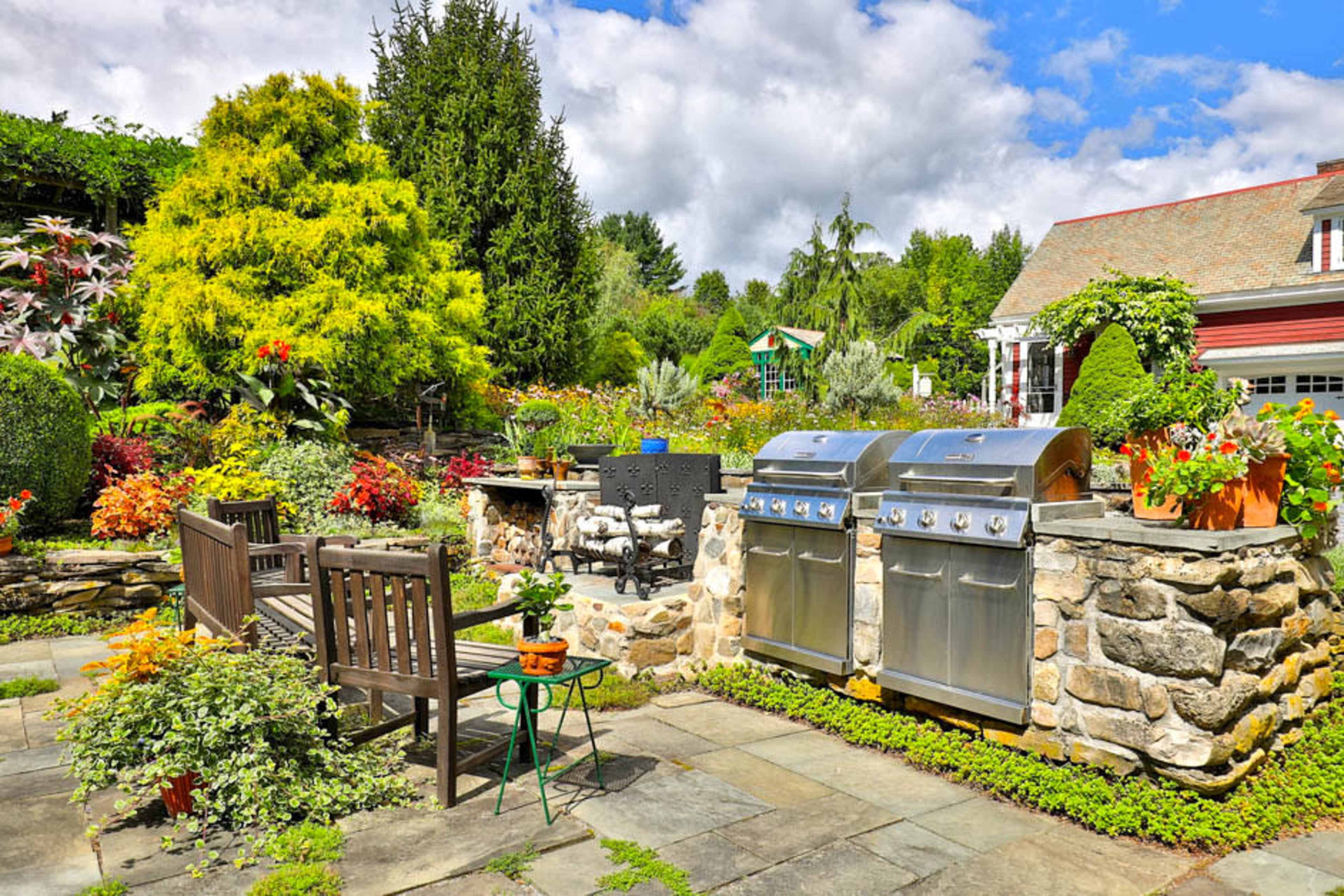 A well-maintained garden with stone grilling stations, wooden benches, and vibrant flowerbeds under a partly cloudy sky.