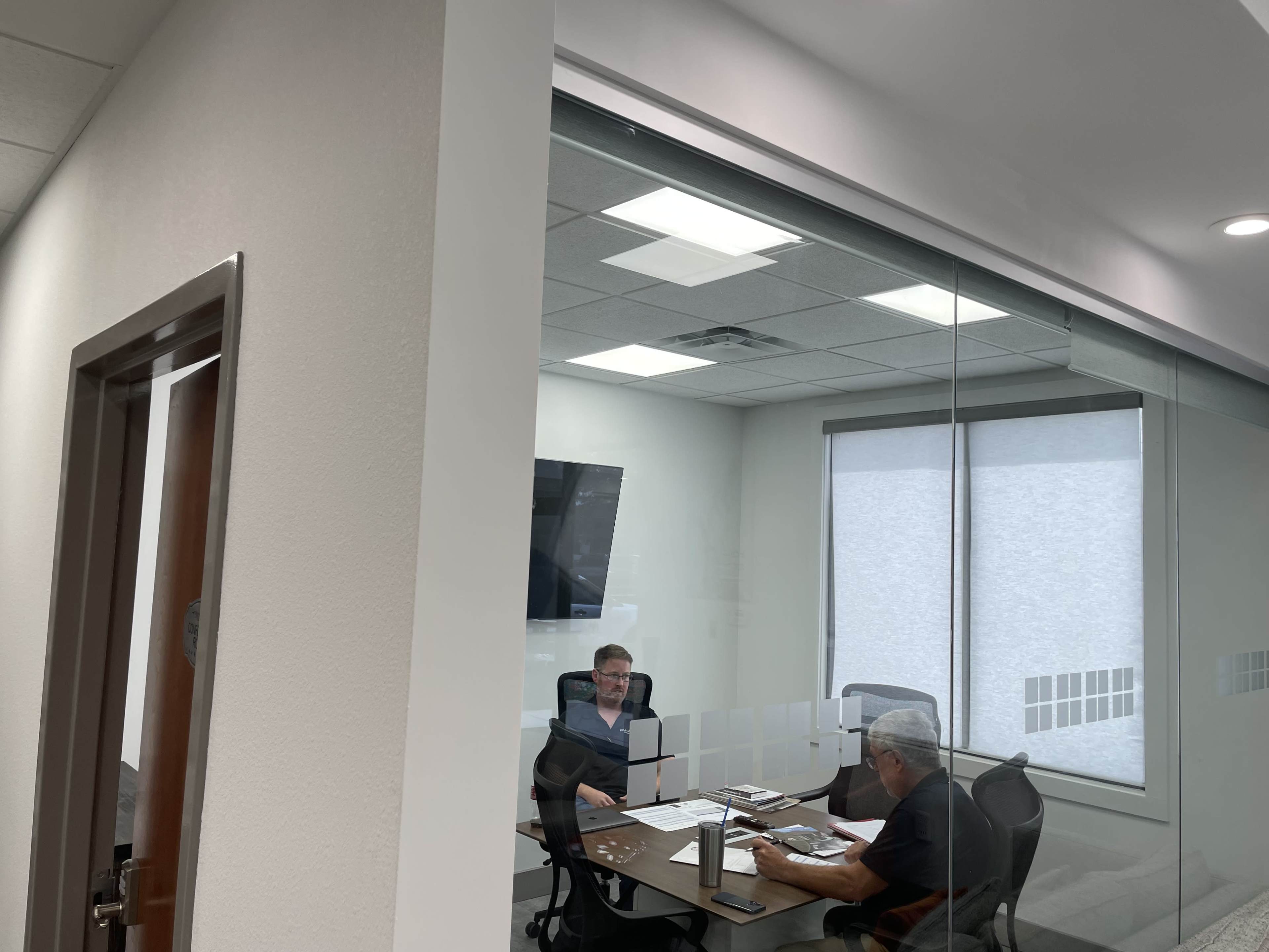 Two men are seated at a table in a conference room, discussing documents while a TV hangs on the wall.