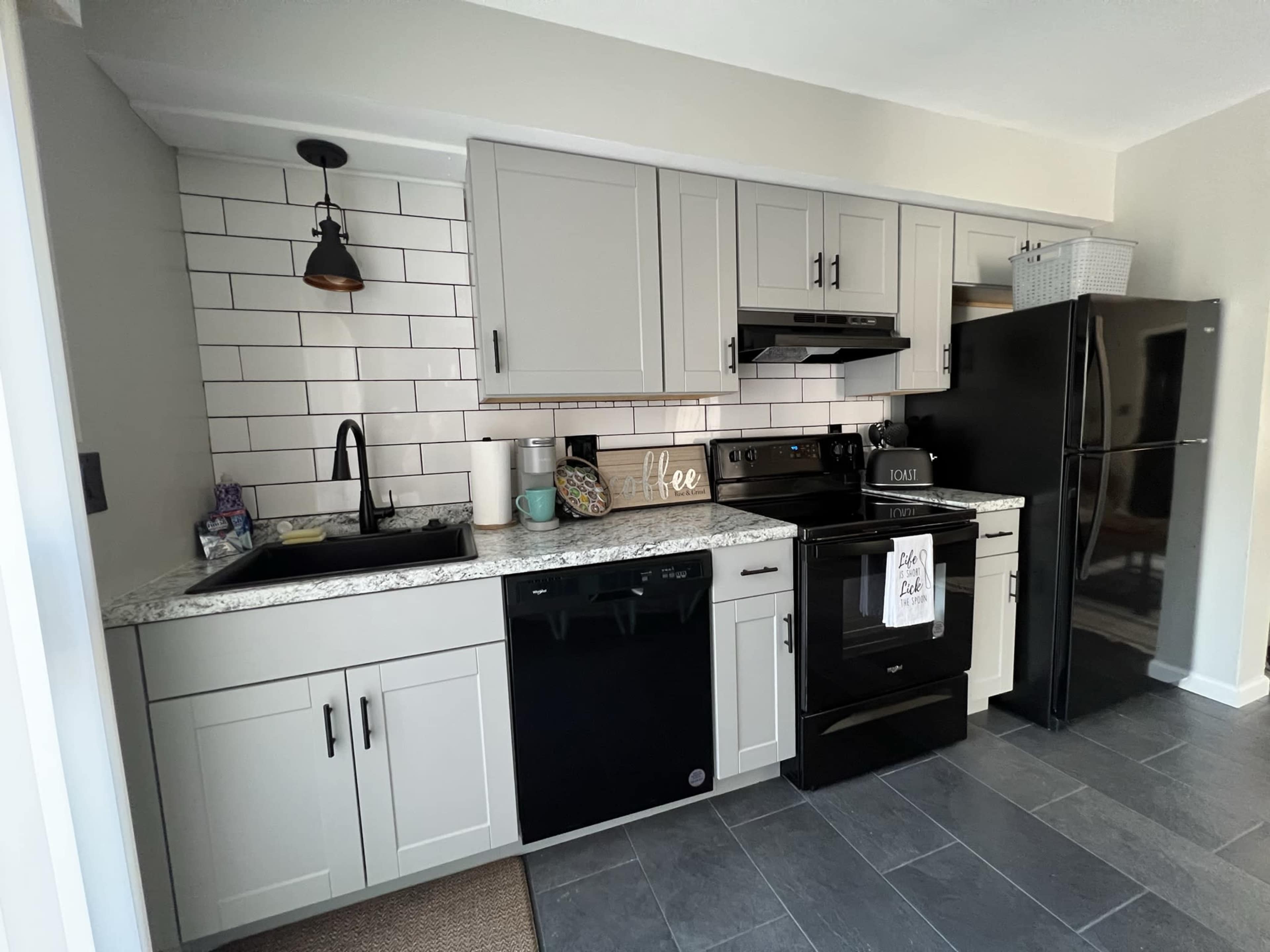 A modern kitchen with gray cabinetry, a black refrigerator, and dark appliances, featuring a white tile backsplash and granite countertops.