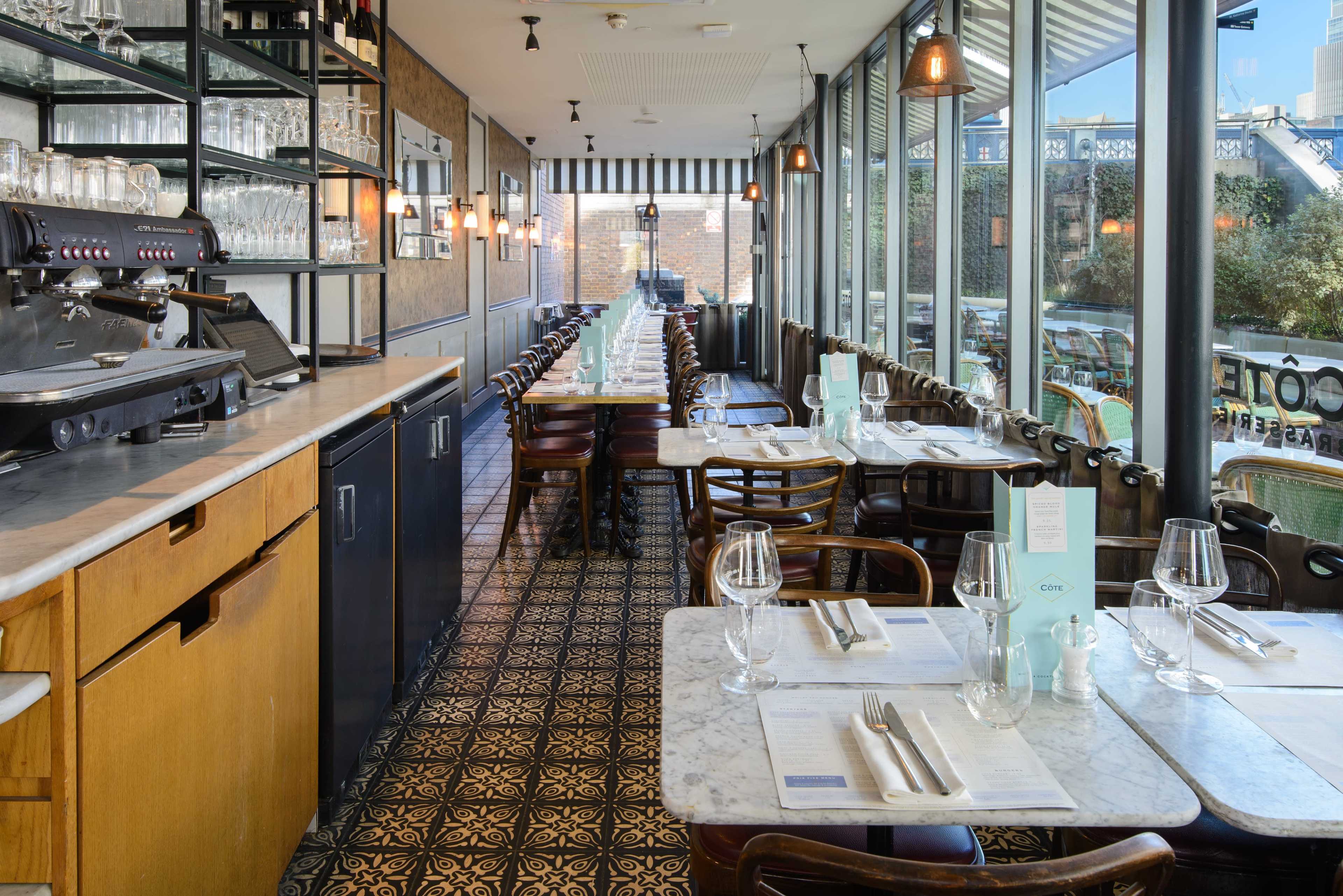 The image shows the interior of a restaurant featuring wooden tables, glassware, and a coffee machine along one wall, with large windows allowing natural light to enter.