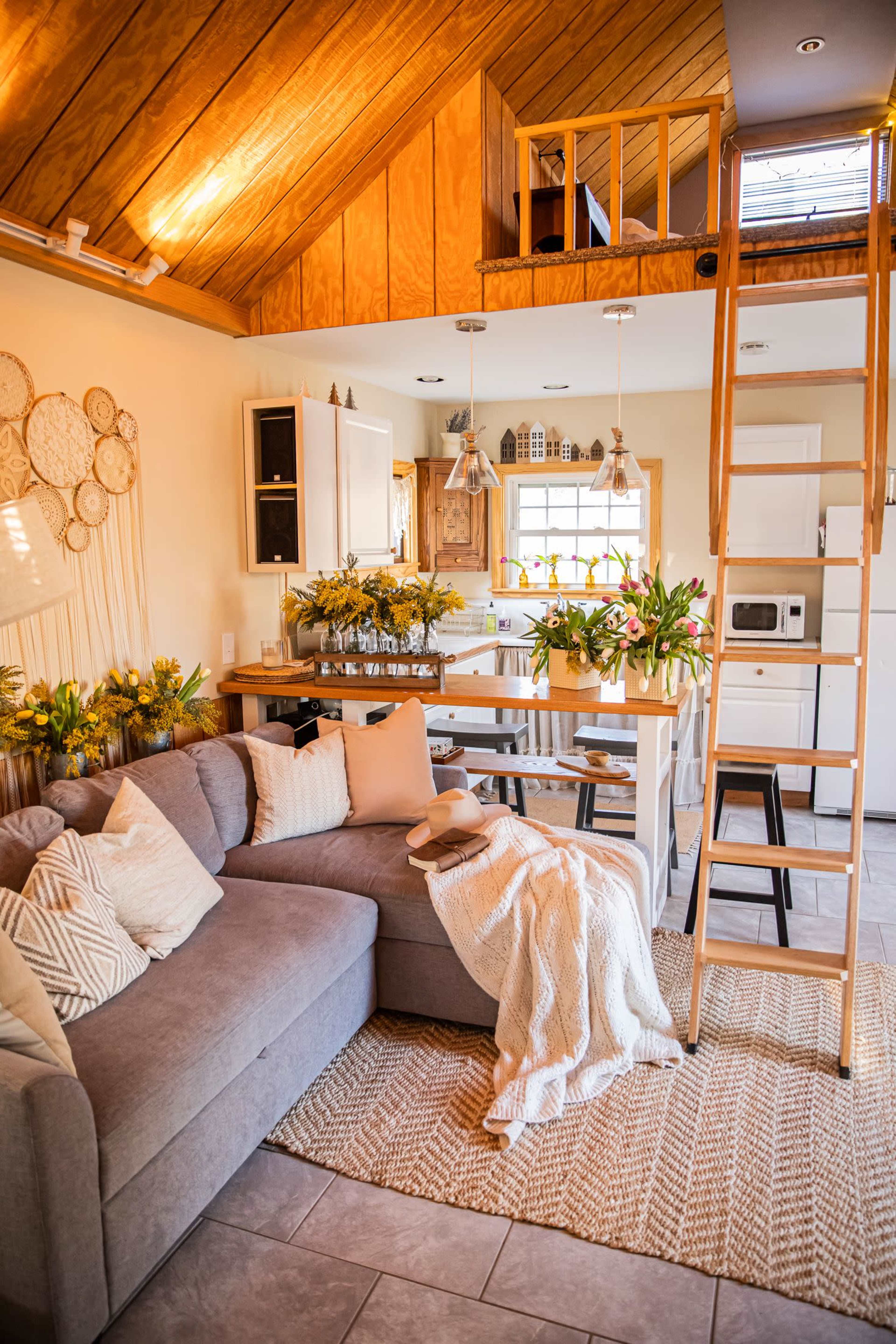 A cozy living space features a gray sectional sofa, a wooden dining table adorned with flowers, and a ladder leading to a loft area.