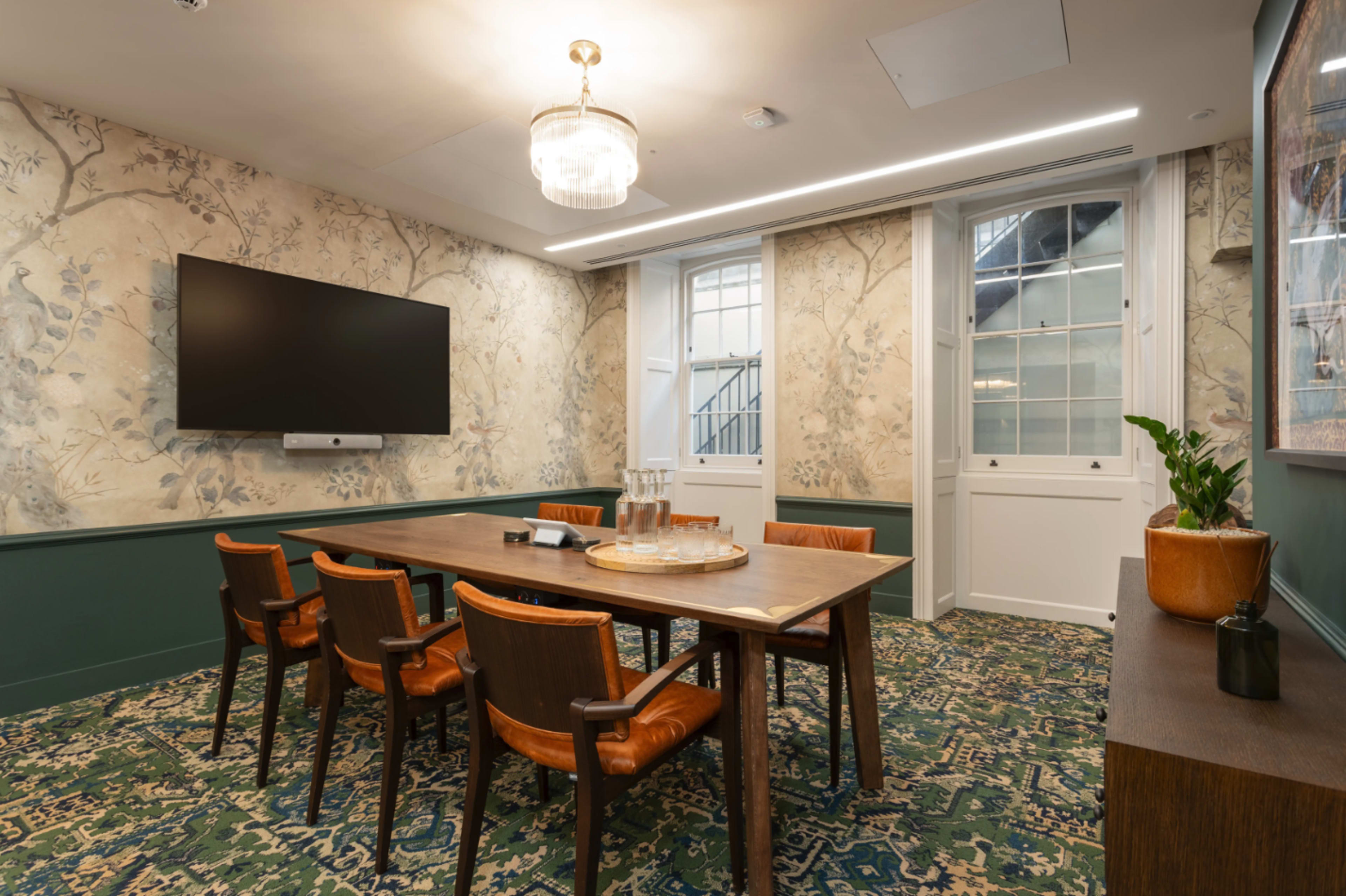 The image shows a small meeting room with a wooden table, six chairs, a large wall-mounted screen, and decorative wallpaper featuring a floral pattern.
