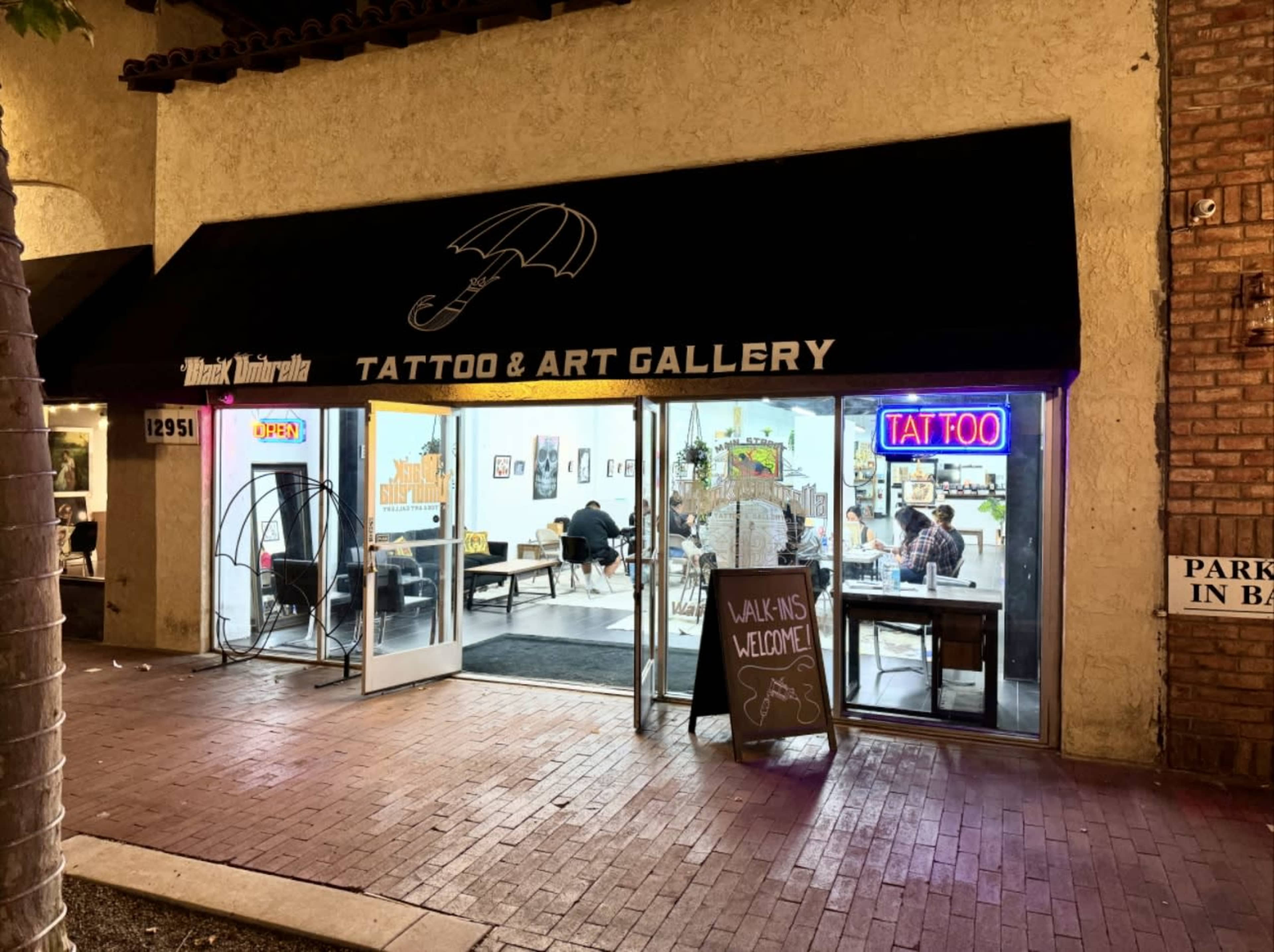 The image shows a tattoo and art gallery storefront with a black awning, featuring a bright sign that reads "TATTOO & ART GALLERY" and an open door welcoming walk-ins.