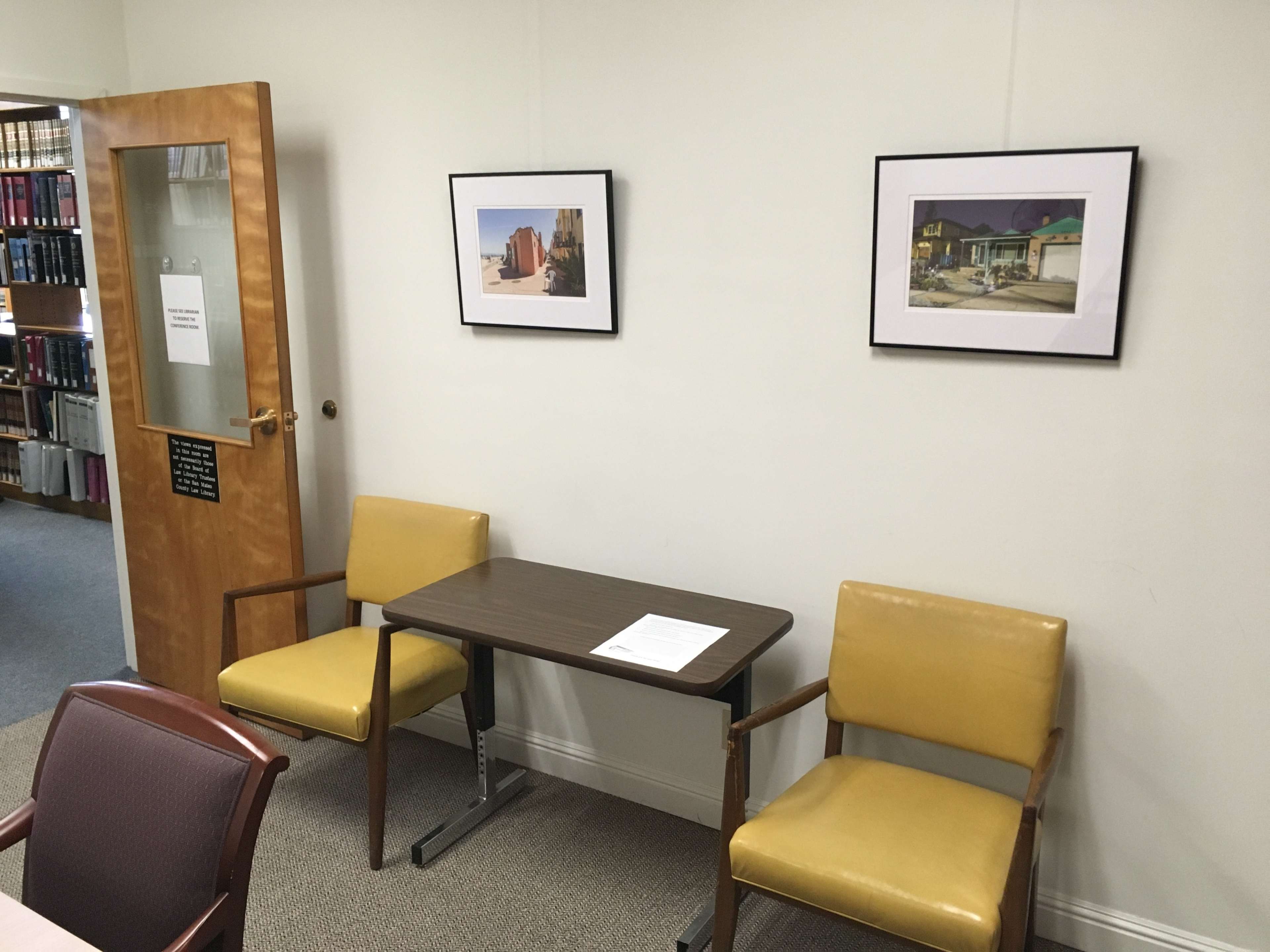 A small room with two yellow chairs, a wooden table, and two framed pictures hanging on the wall.
