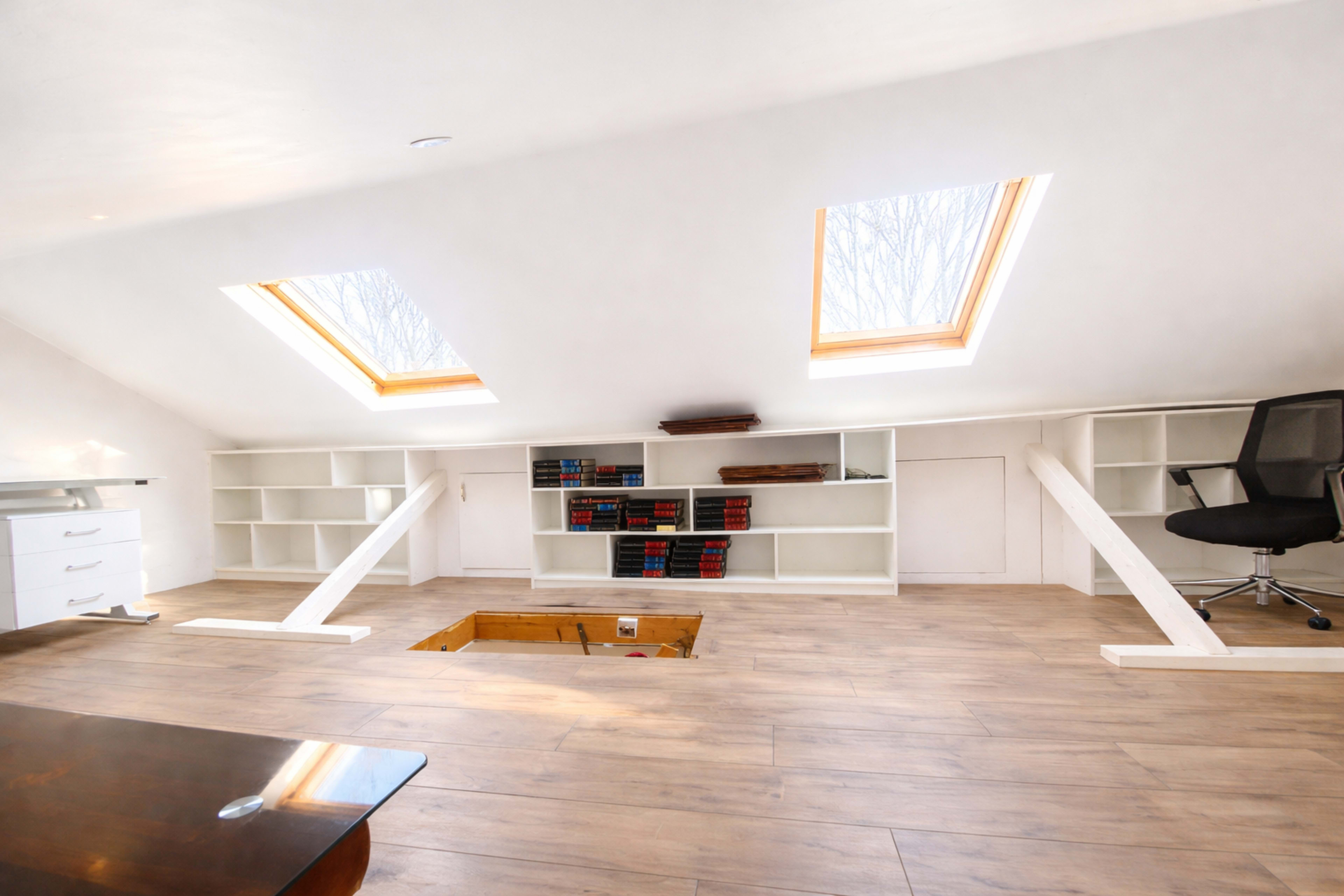 The image shows a well-lit attic space featuring two skylights, wooden flooring, built-in shelves with books, and a small opening in the floor.