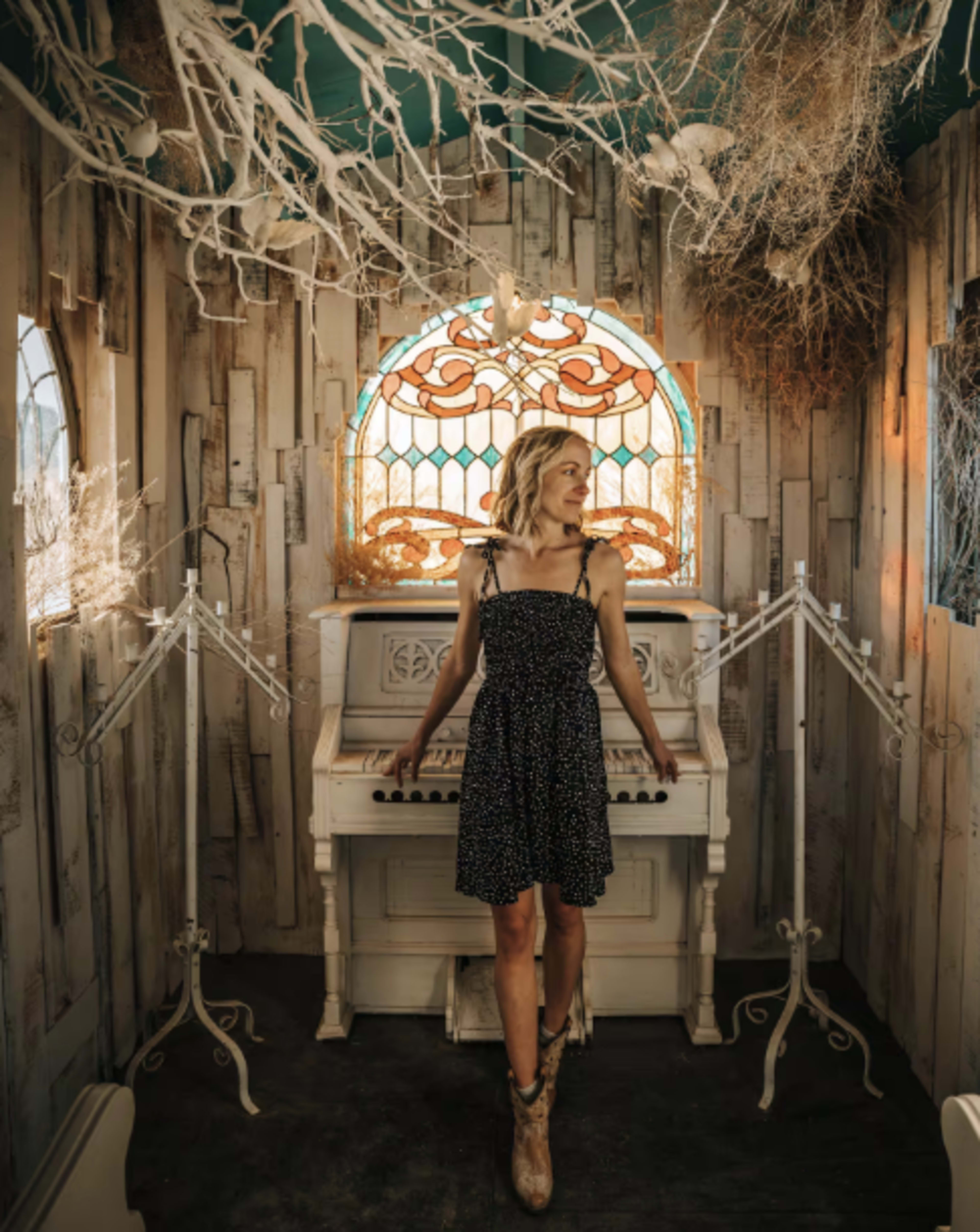 A woman in a black polka-dot dress stands by a white piano in a room adorned with decorative branches and a stained glass window.
