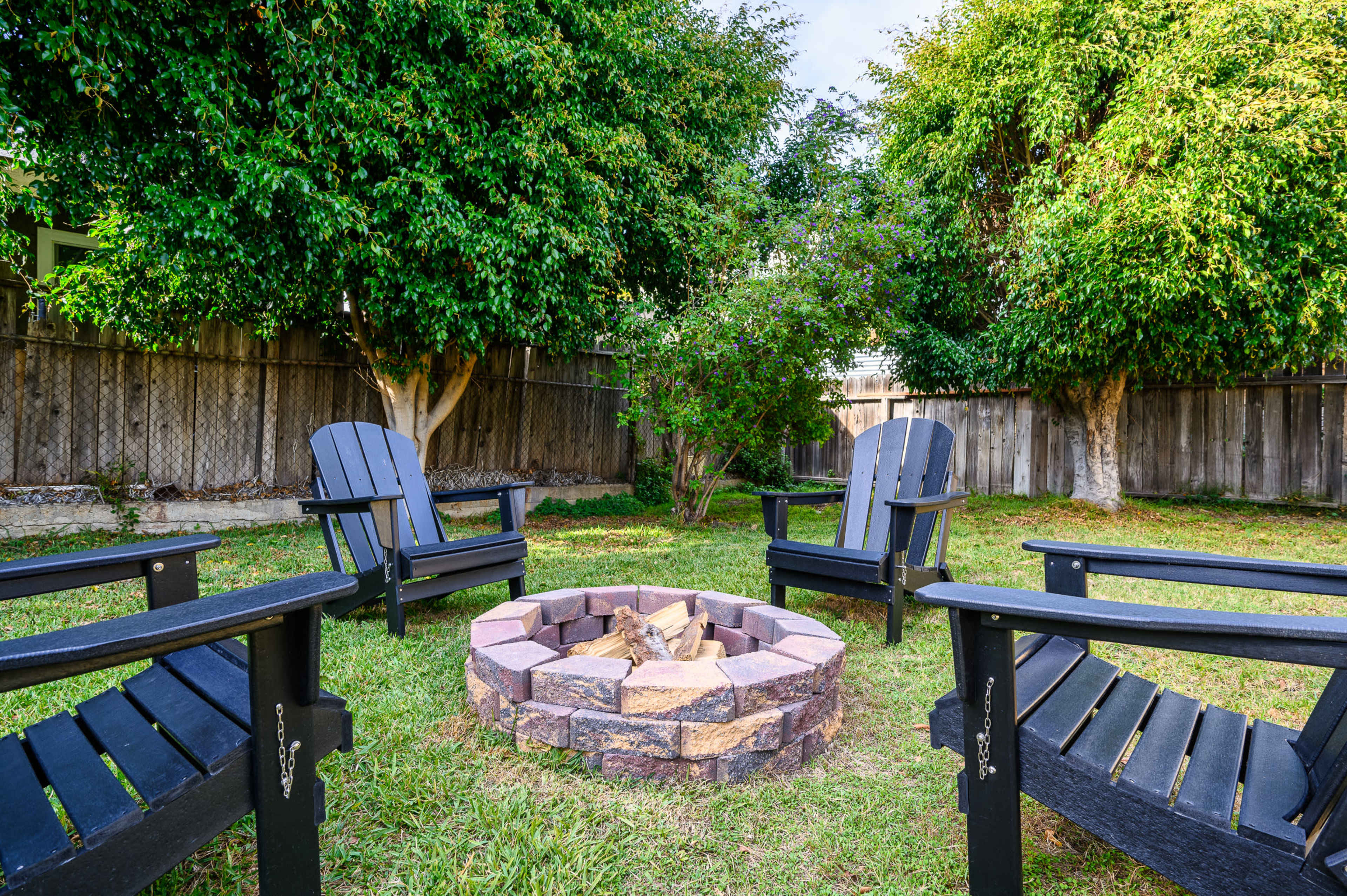 The image shows a backyard with four black Adirondack chairs arranged around a circular stone fire pit, surrounded by green grass and trees.