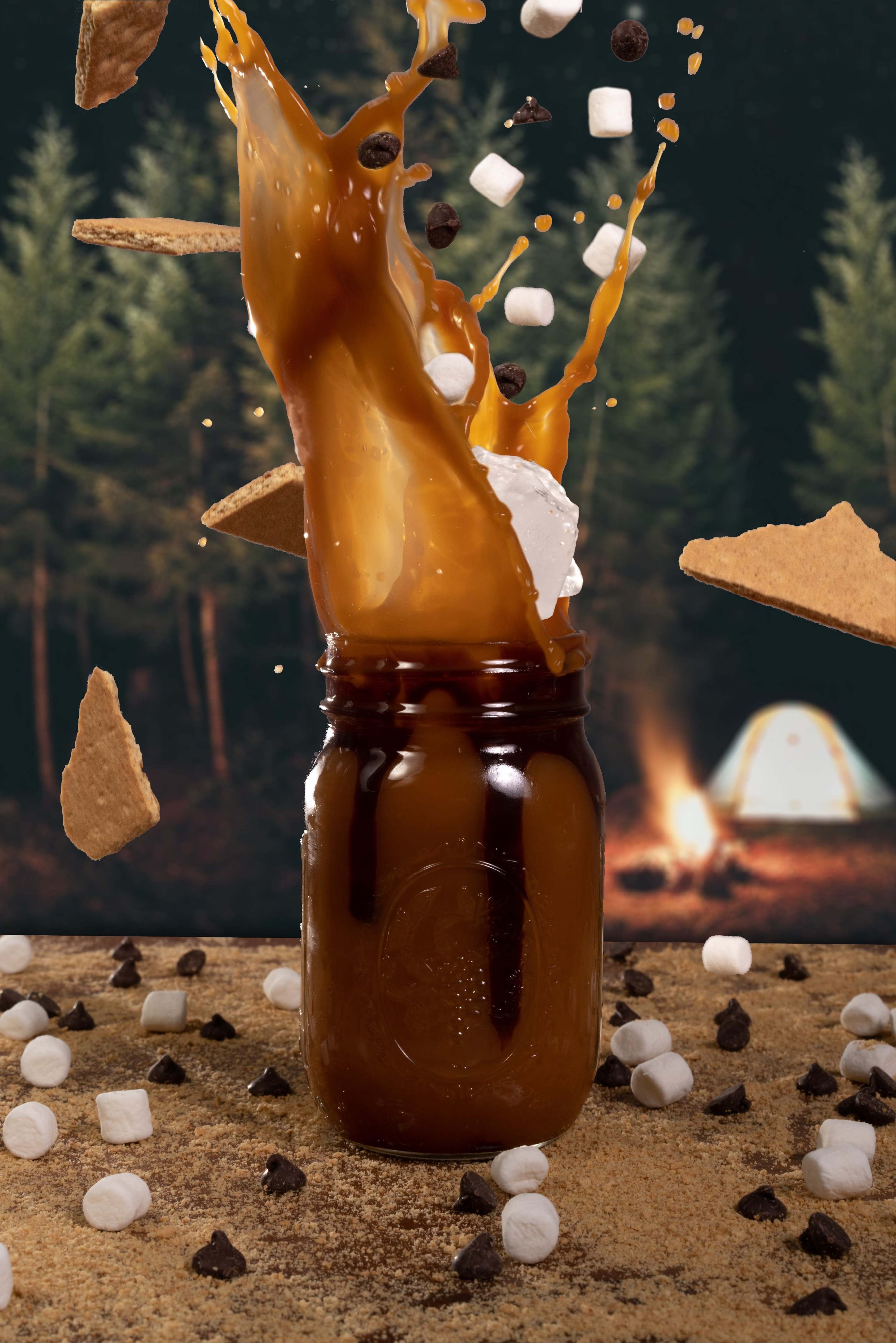A jar filled with a liquid is splashing while surrounded by scattered graham crackers, marshmallows, and chocolate chips in a forest campsite setting.