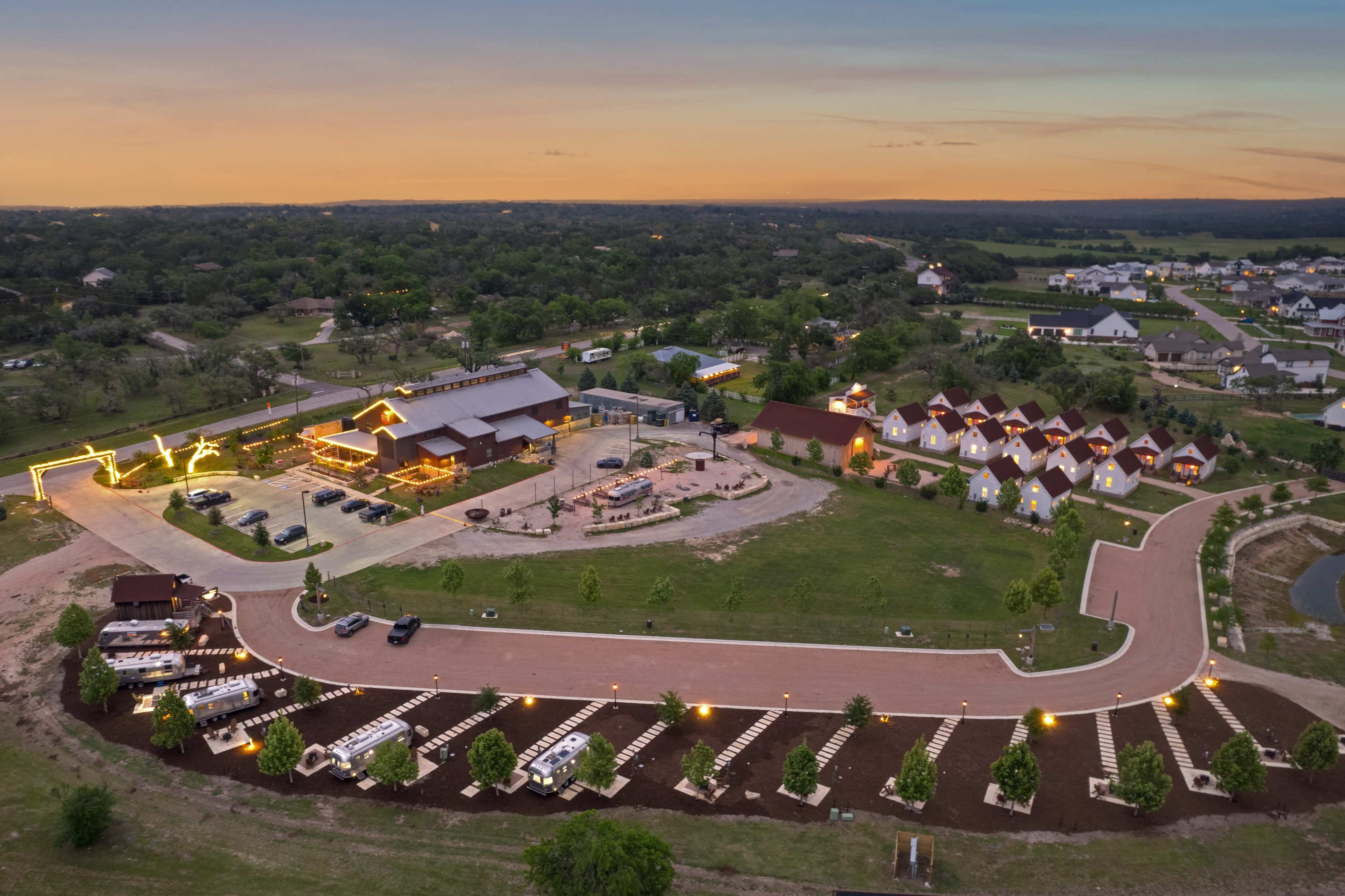 The image shows a large outdoor recreational facility with cabins, parking areas, and landscaped grounds, illuminated by lights during dusk.