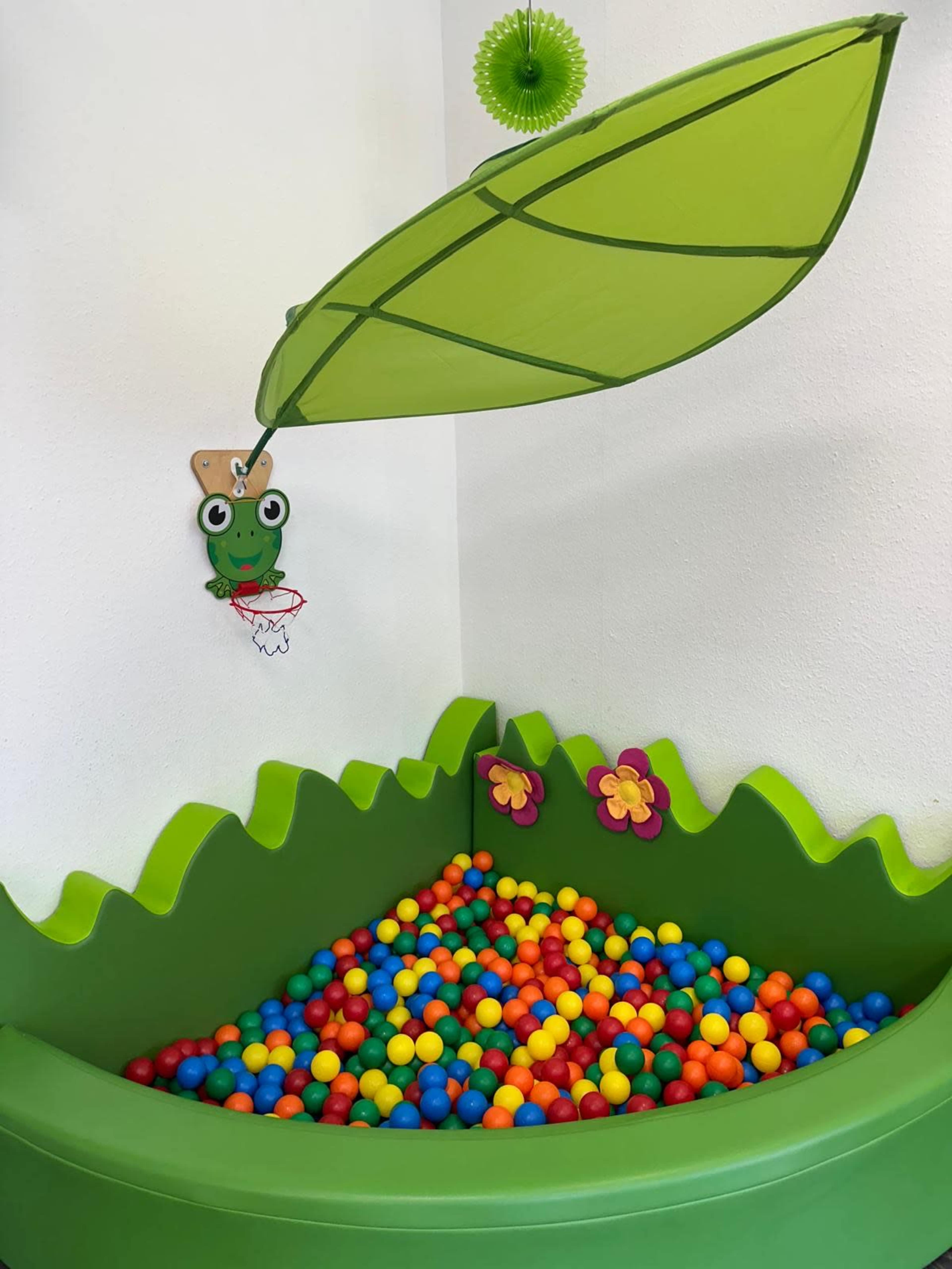 The image shows a colorful ball pit filled with red, blue, yellow, and green balls, surrounded by a green, grassy-shaped border and decorated with flowers and a hanging frog toy.
