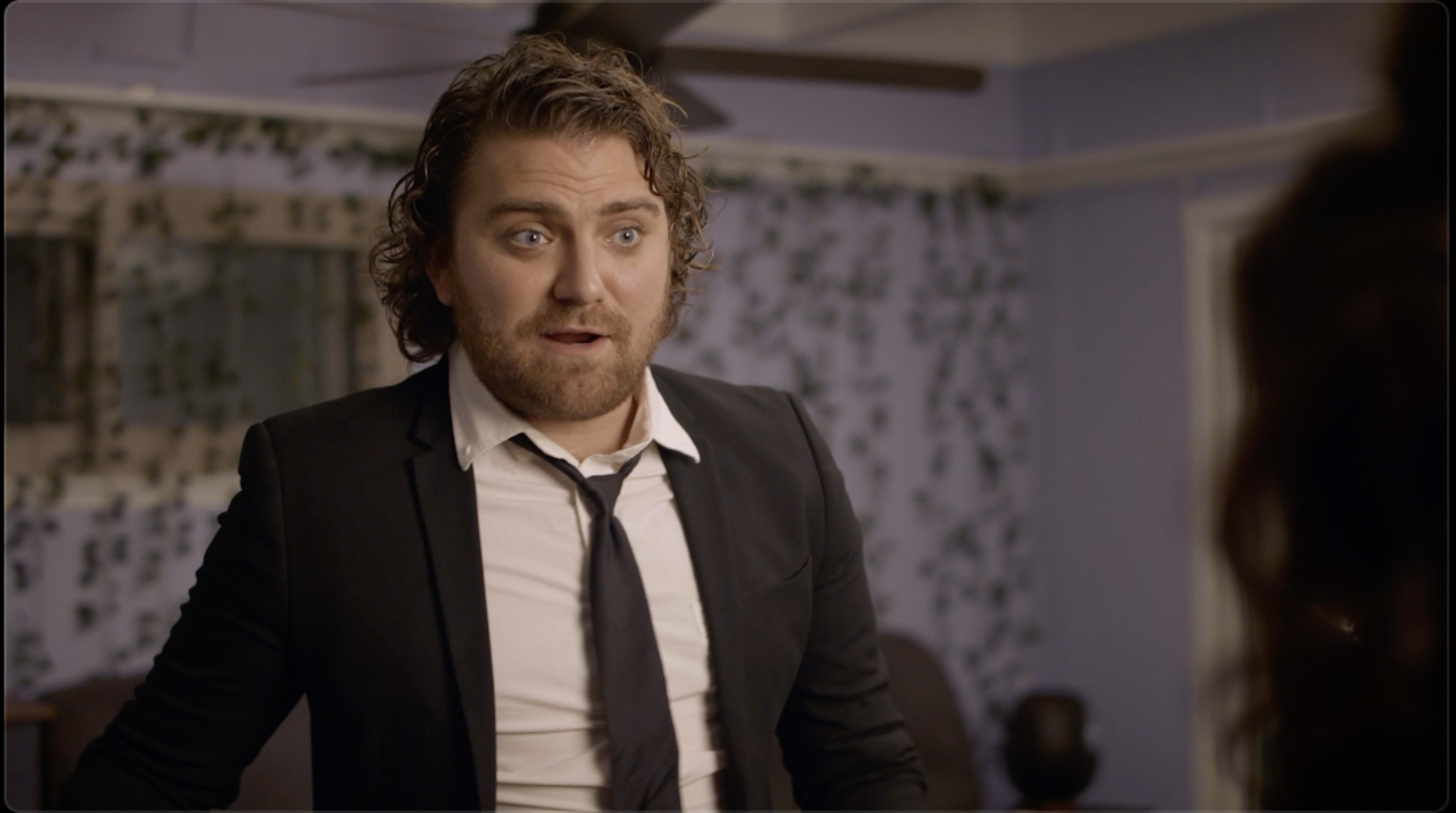 A man in a suit with disheveled hair stands in a room with floral wallpaper, looking surprised.