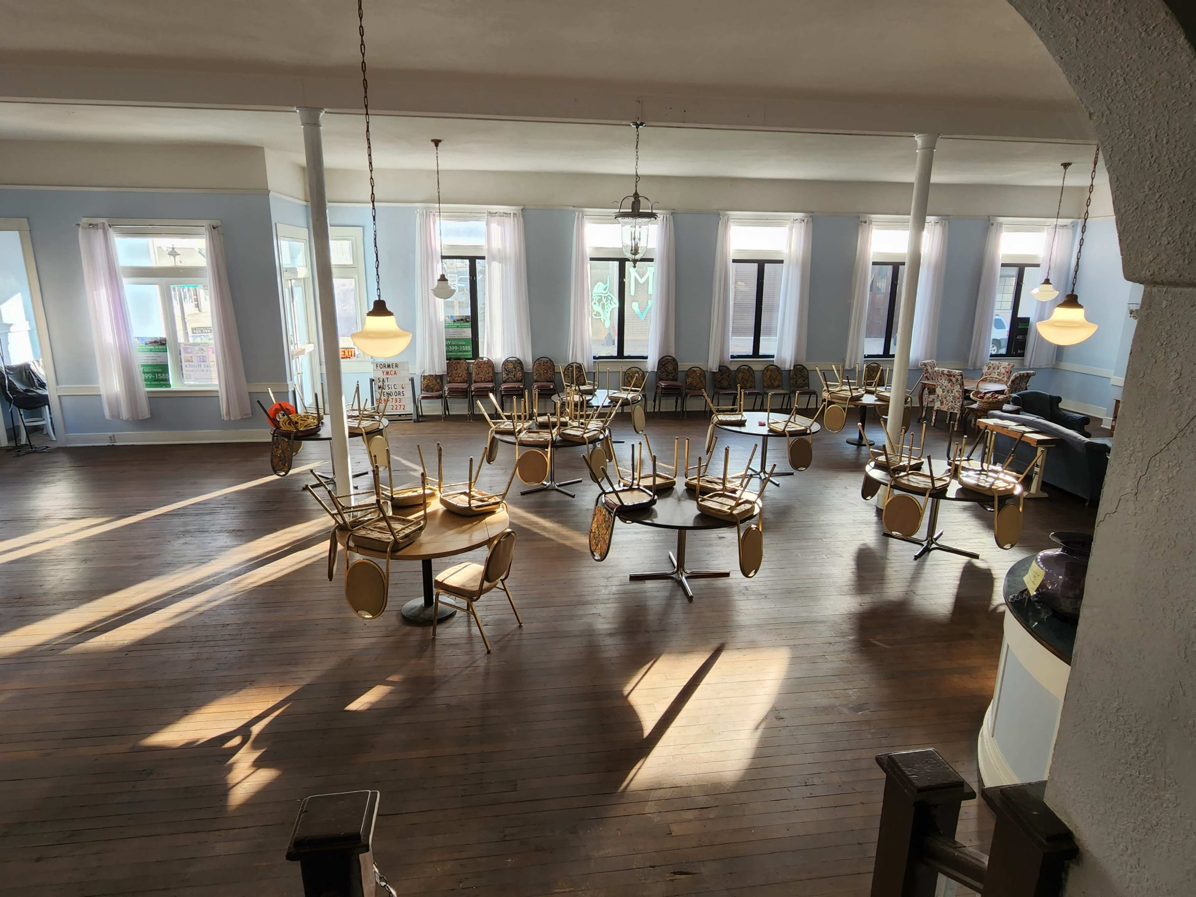 A spacious dining area with tables and chairs stacked upside down on the wooden floor, illuminated by natural light streaming through large windows.