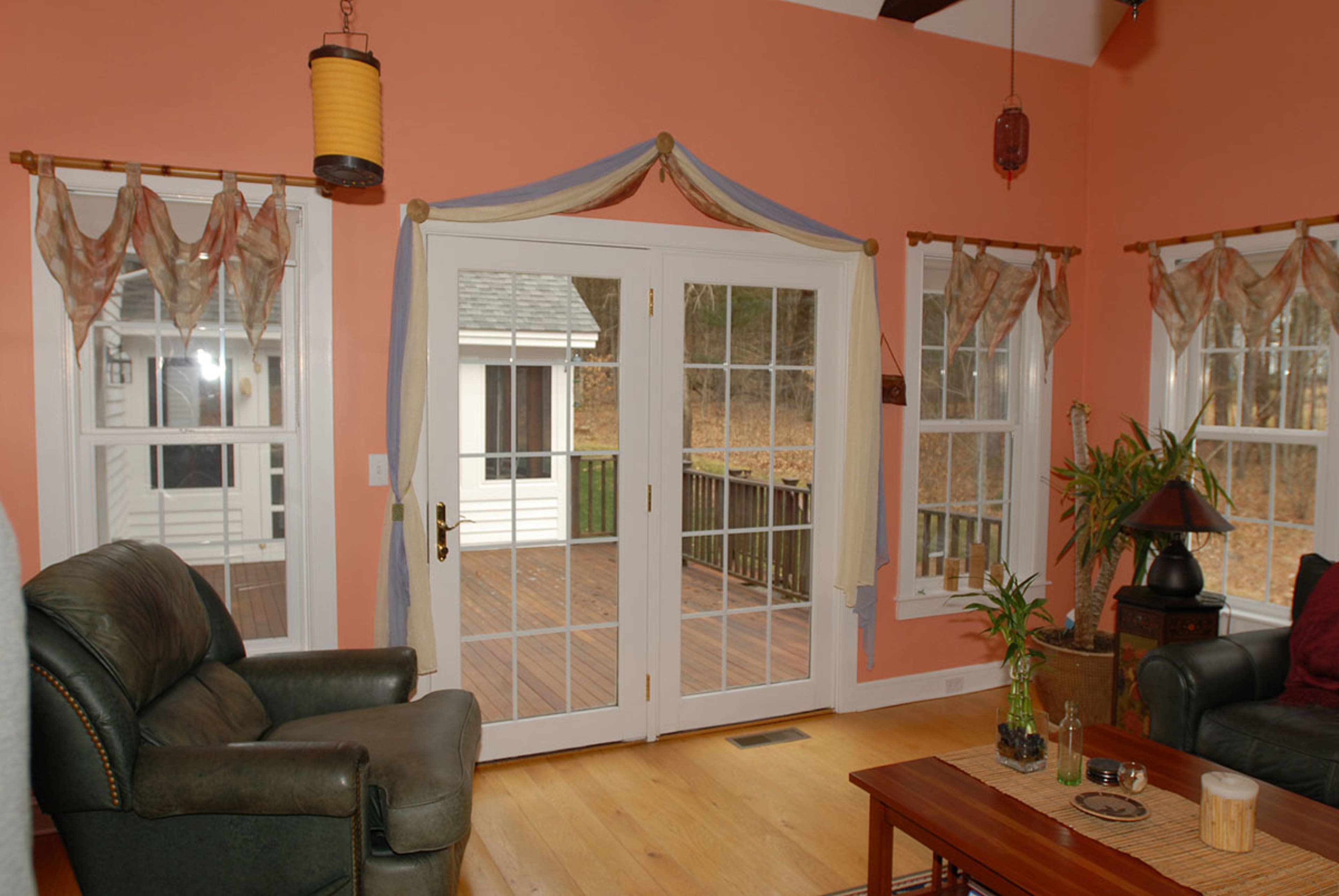The image shows a cozy living room with a pair of French doors leading to a deck, flanked by windows draped with curtains.